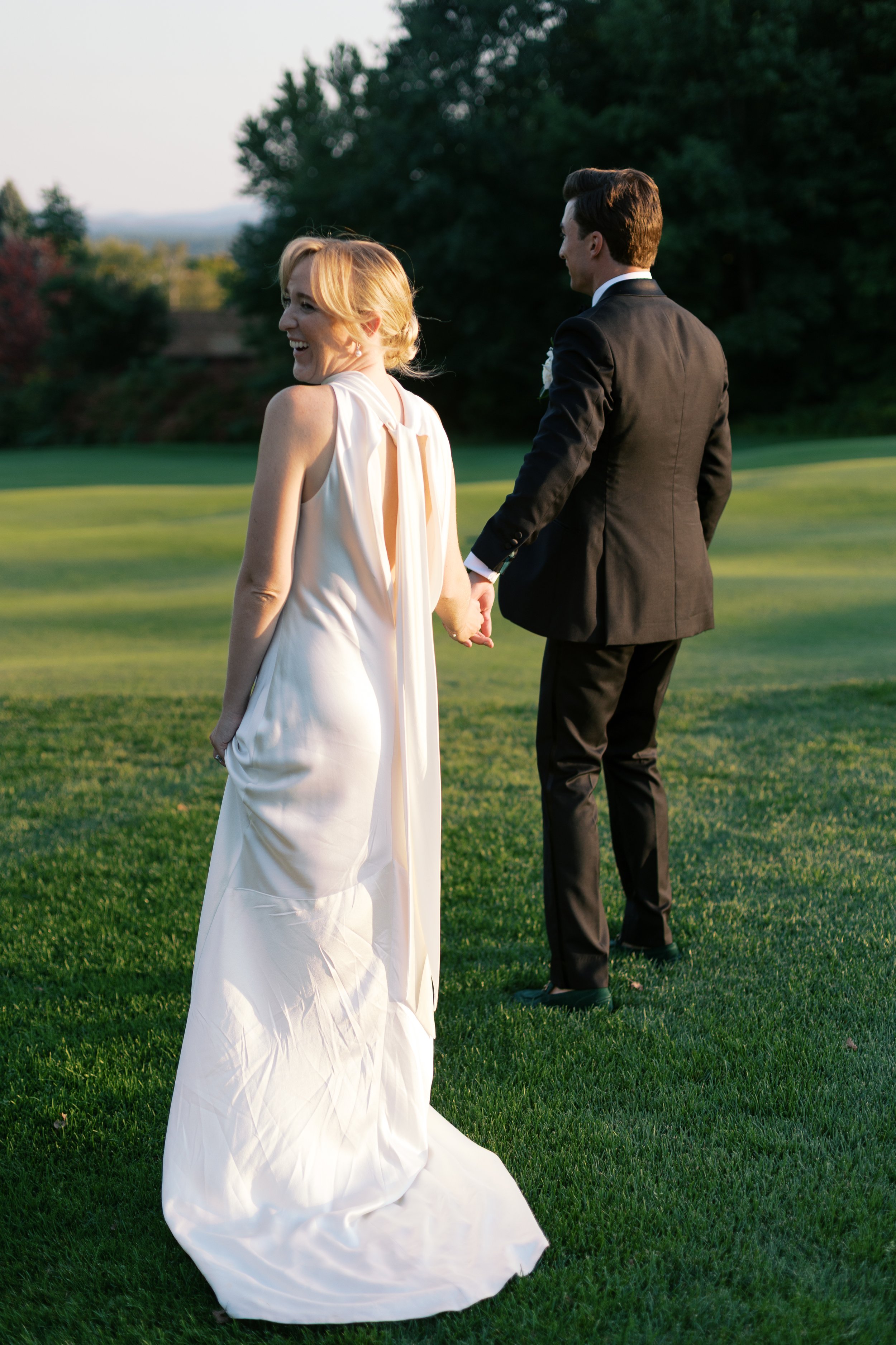 A bride and groom holding hands and walking on a green lawn during sunset at their outdoor wedding.