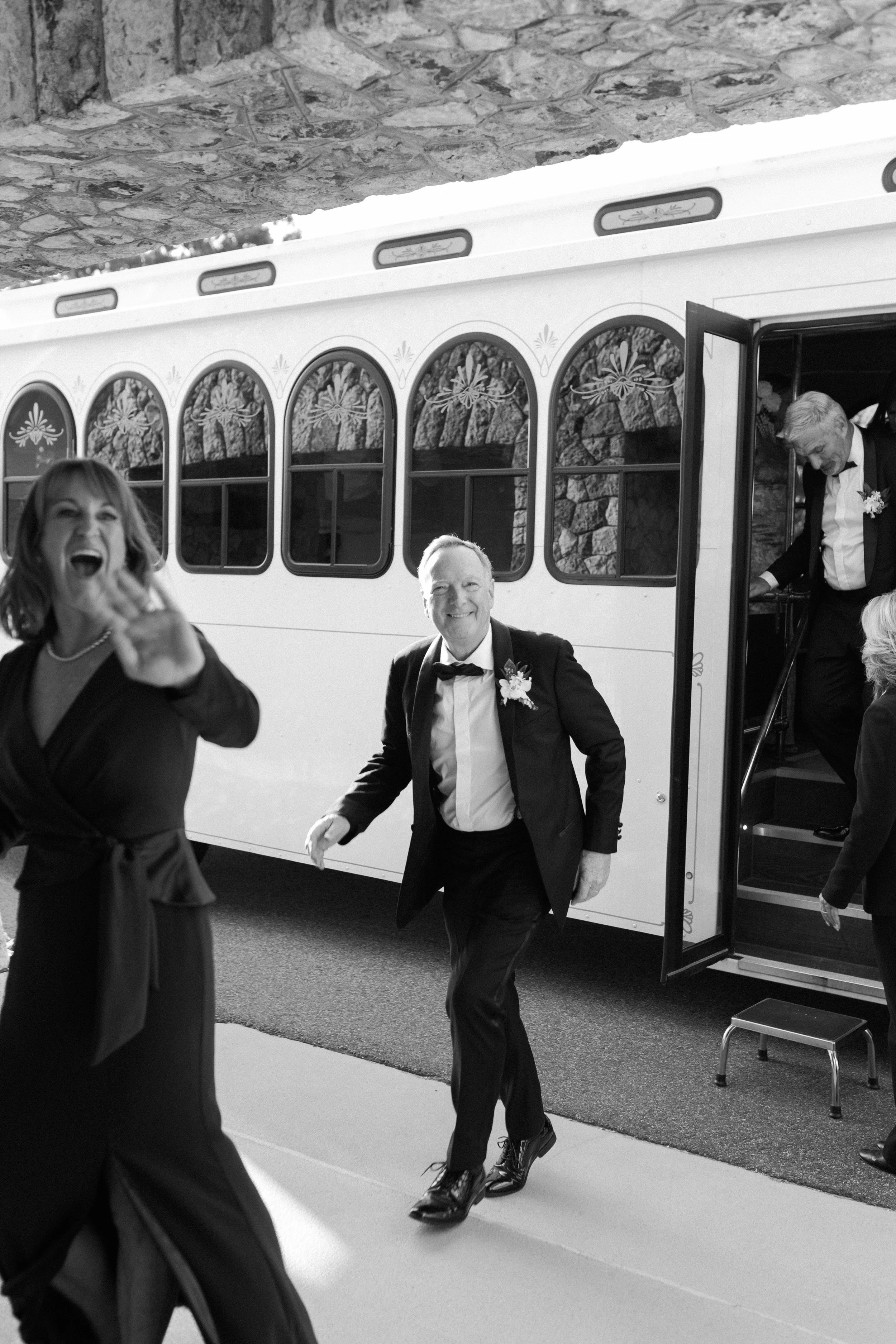 Black and white photo of a man and a woman dressed in formal attire celebrating outside near a bus. The woman is smiling and waving, and the man is walking towards the camera with a cheerful expression. The bus has decorative windows and is parked un