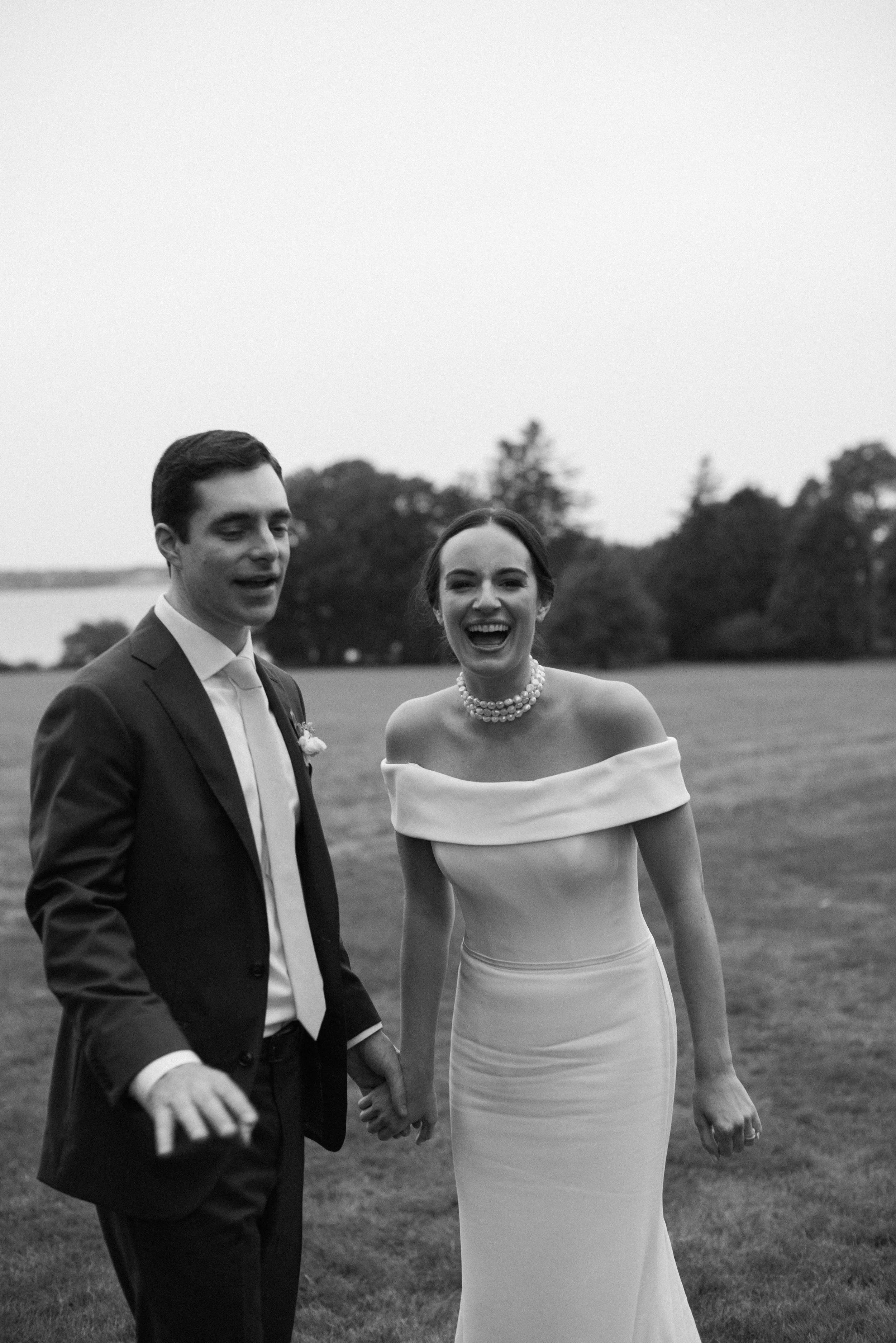 A black-and-white photograph of a happy couple, woman in an off-the-shoulder wedding dress with pearl necklace, man in a suit, holding hands outdoors in a grassy area with trees in the background.