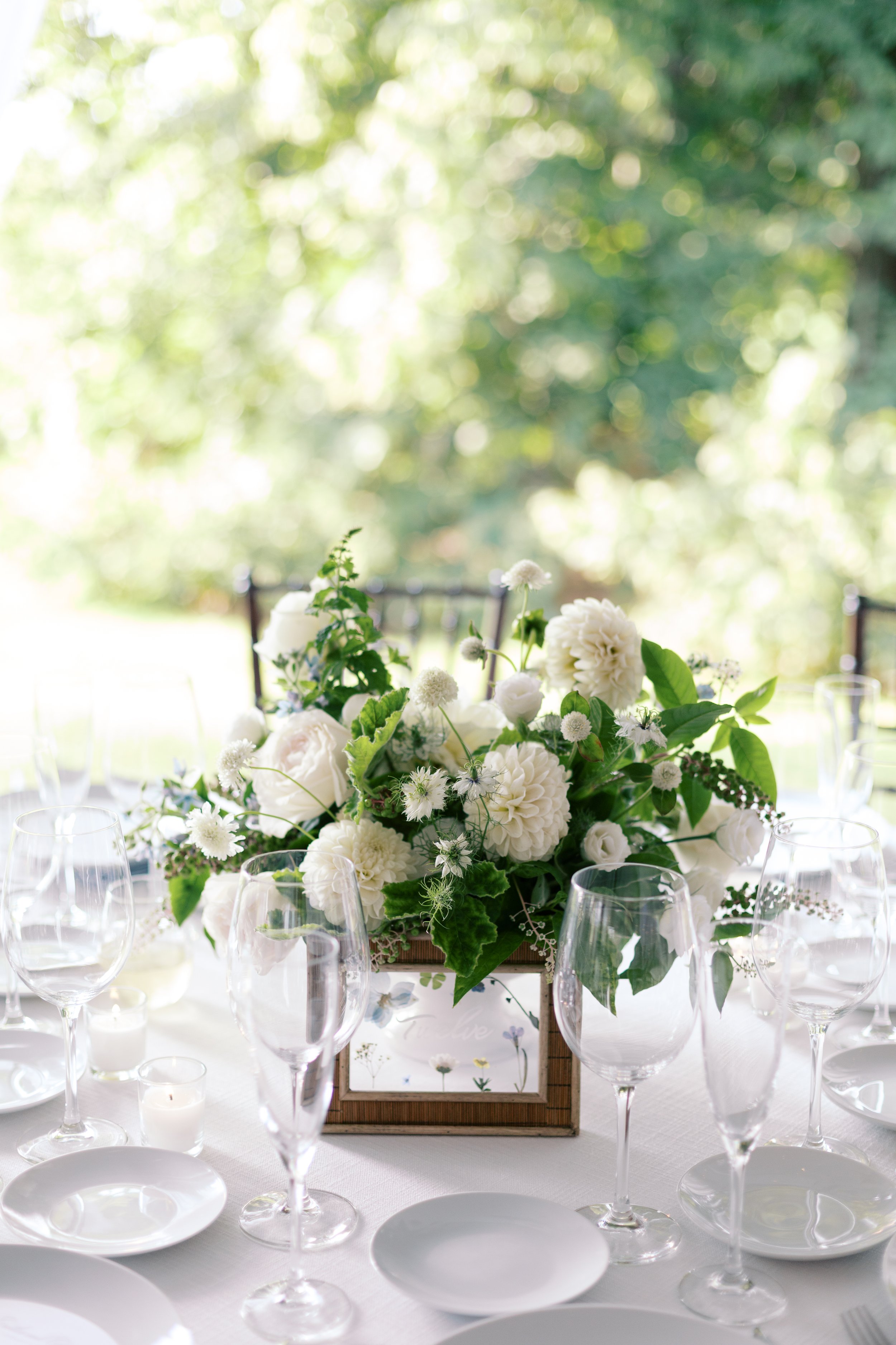 A table centerpiece with white flowers and green leaves, surrounded by empty wine glasses and small candles, set outdoors with a blurred green background.
