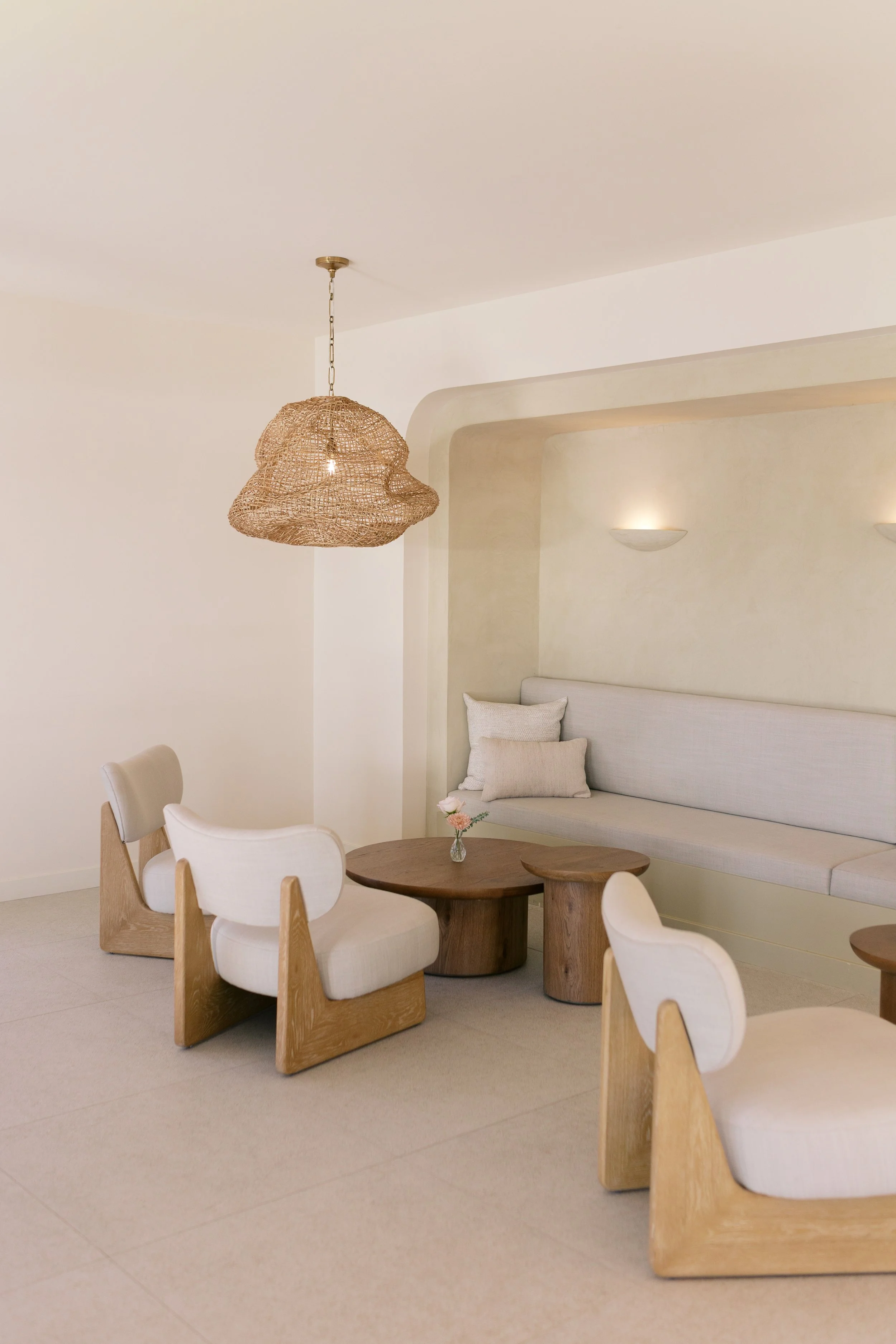 Minimalist living room with beige chairs, wooden tables, a built-in bench with pillows, wall-mounted lights, and a woven pendant lamp.