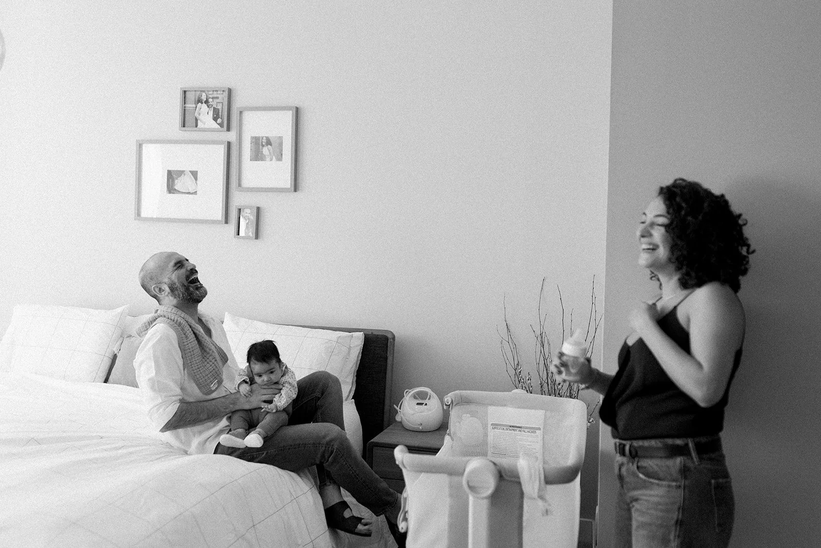 Black-and-white photo of a joyful family moment in a bedroom. An elderly man sitting on a bed holding a small child, both laughing. A woman standing nearby, also laughing, with her hand on her chest. The room has a nightstand with a small device and 