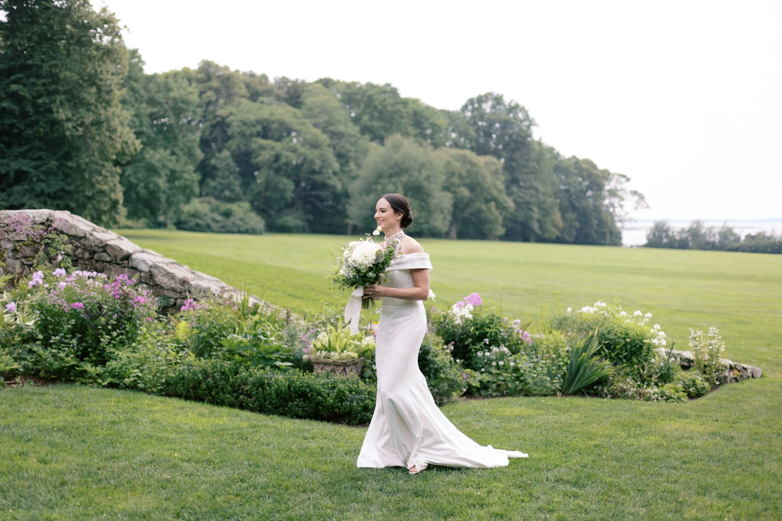 A bride in a white wedding gown holding a bouquet of white flowers standing on a lush green lawn with a garden and trees in the background.