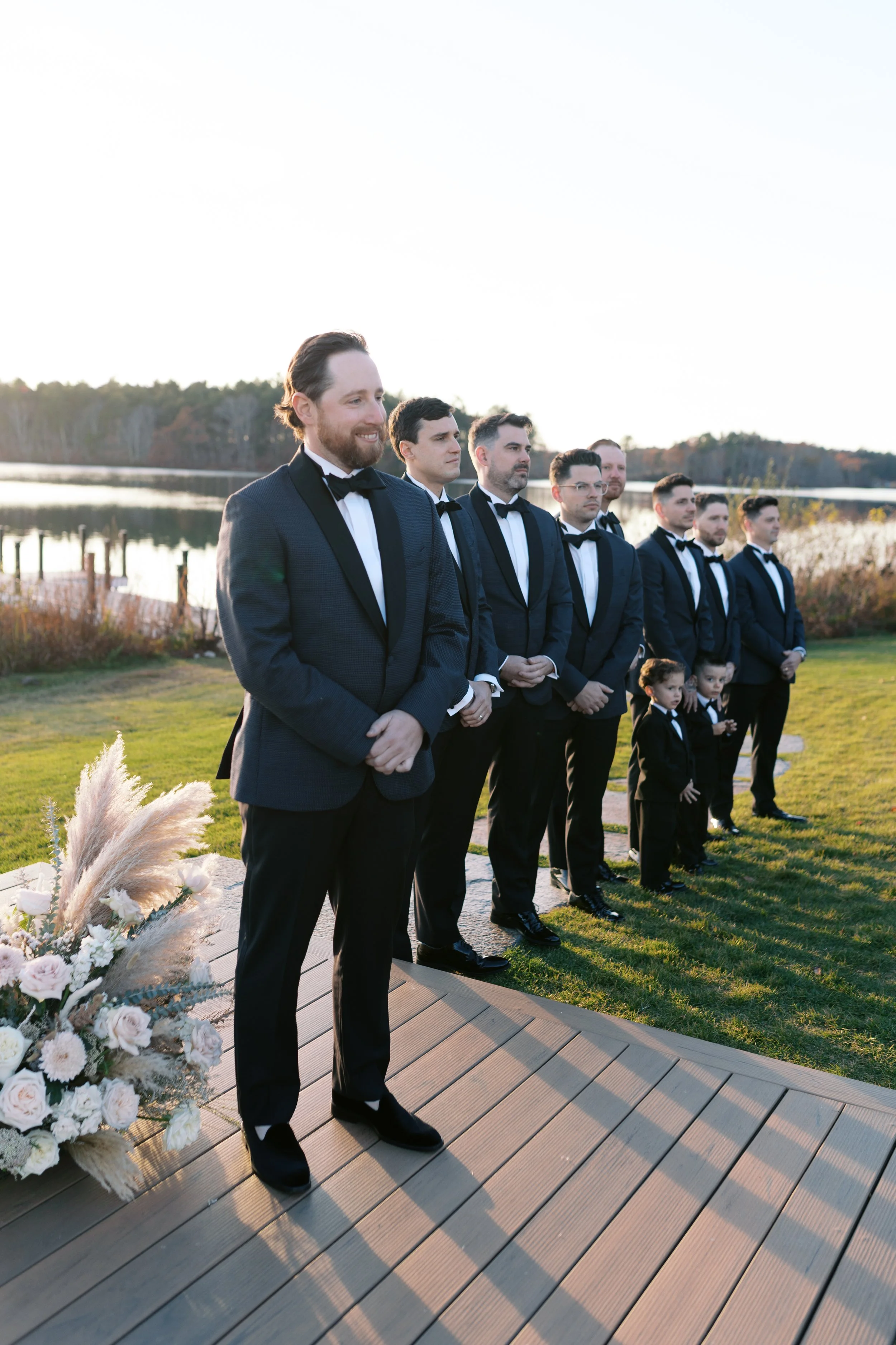 Groom and groomsmen in formal suits standing outdoors during a wedding ceremony near a lake with greenery and flowers in the background.