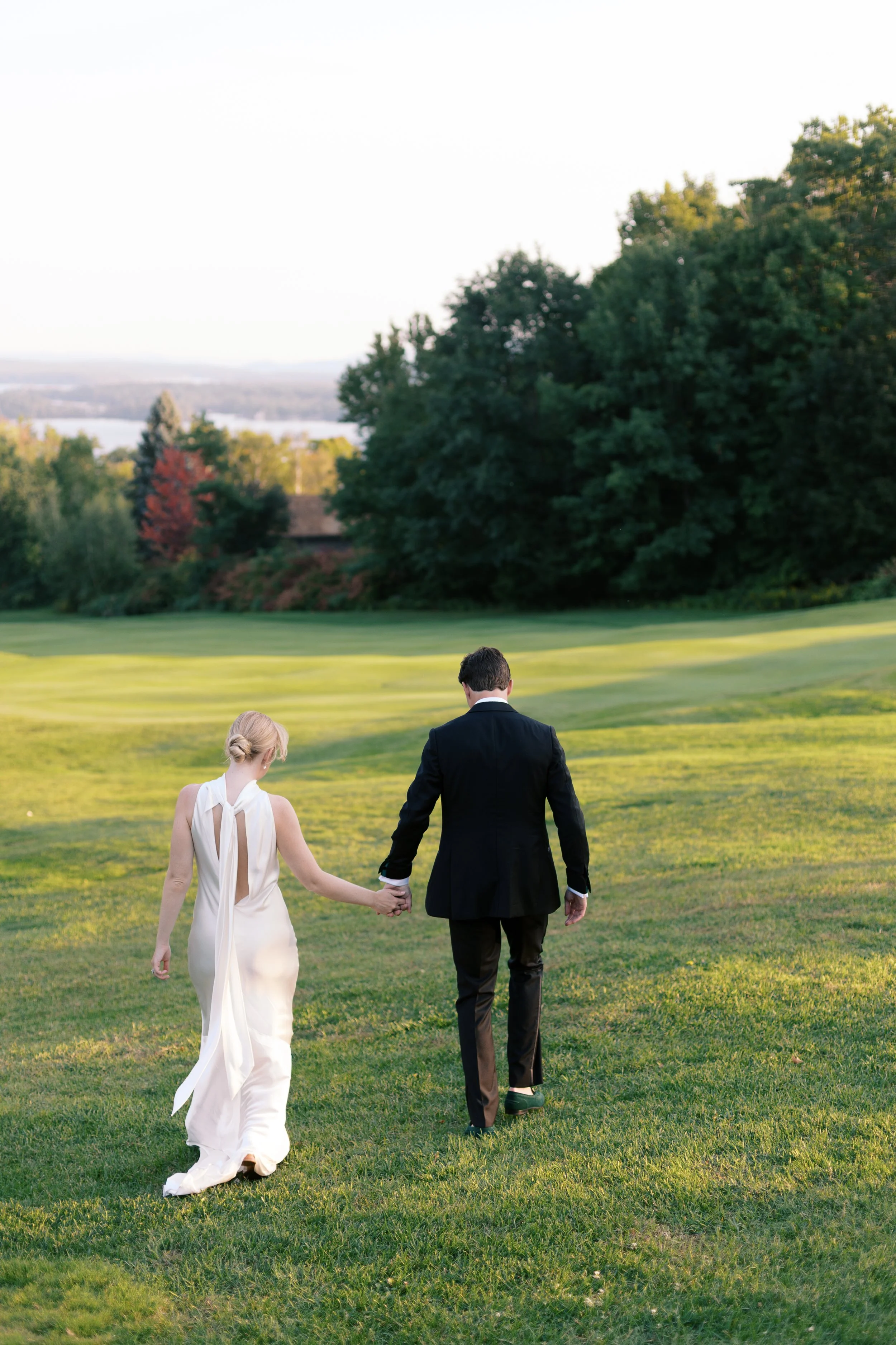 A bride and groom walk hand in hand across a grassy field in a park, with a background of trees and a lake.