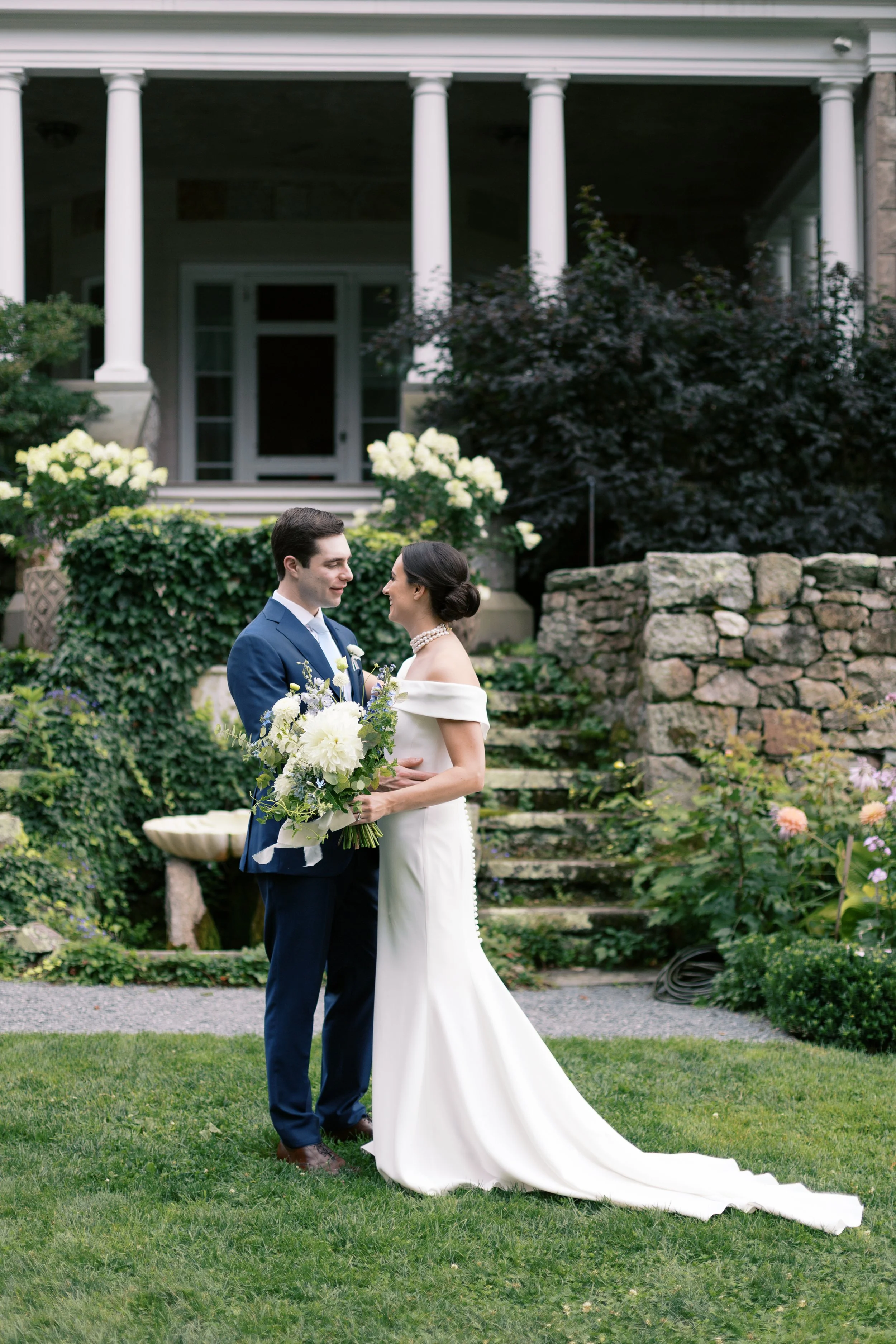 A bride and groom on their wedding day, standing on a lawn with a garden and house in the background, the bride holding a bouquet and wearing a white gown, while the groom is dressed in a blue suit.