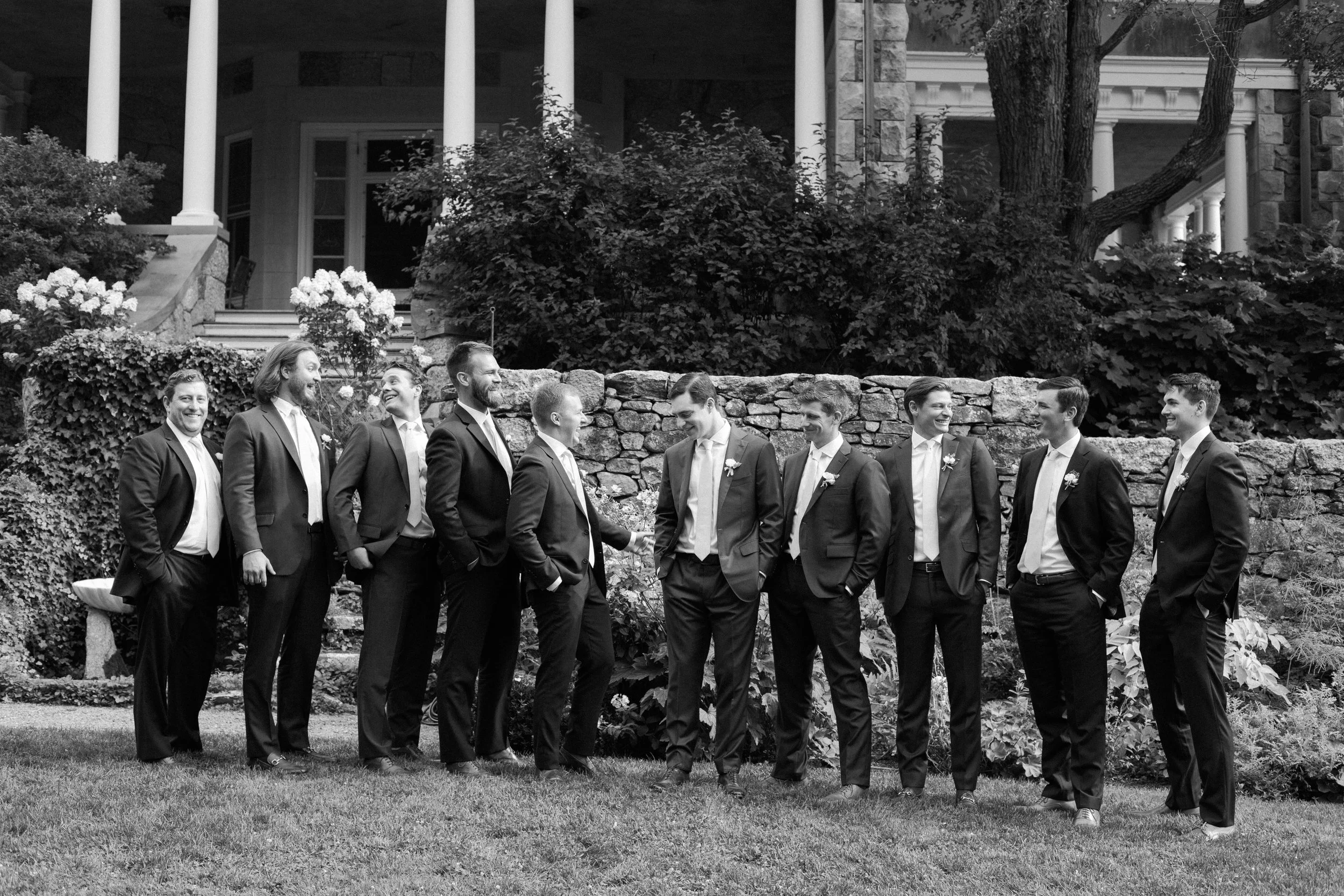 Group of men in suits standing outdoors in front of a stone wall, smiling and engaging in conversation, with a house and trees in the background.