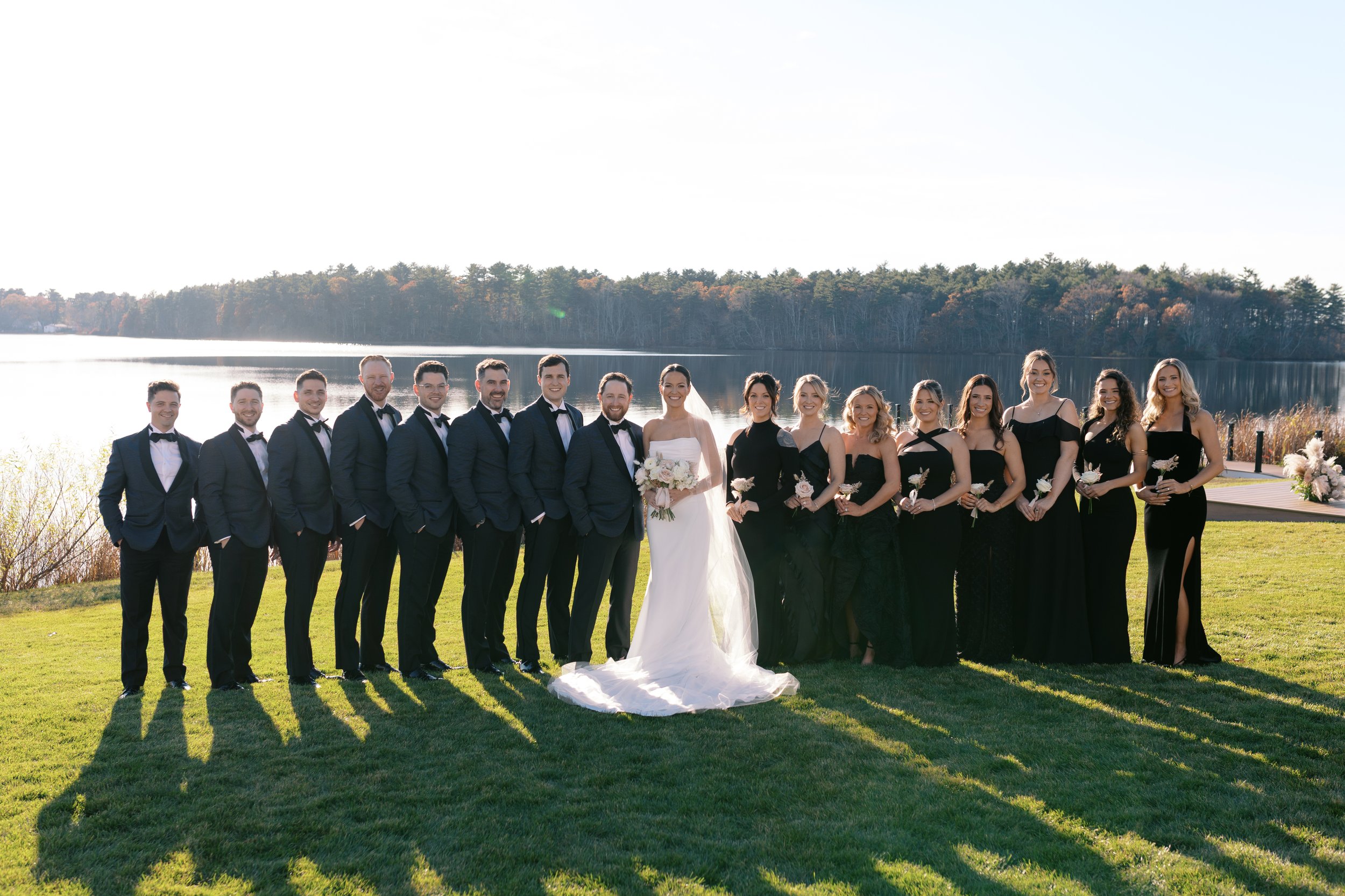 A wedding party standing outdoors on grass near a lake, with a line of trees in the background. The bride is in the center wearing a white gown and holding a bouquet, surrounded by bridesmaids in black dresses and groomsmen in navy suits with bow tie