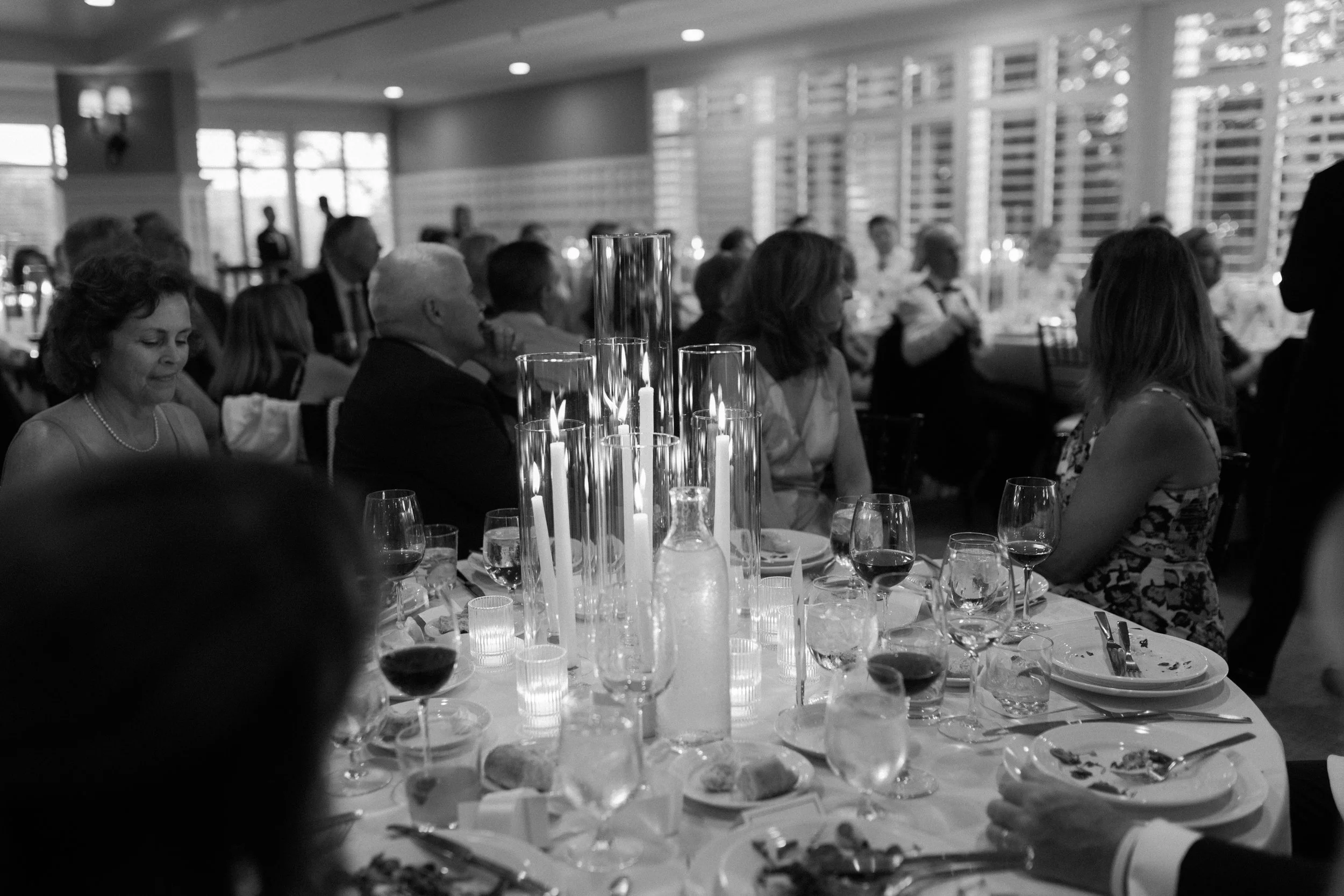 People seated at a decorated dining table during a formal event or celebration, with tall candles in glass holders as centerpieces.