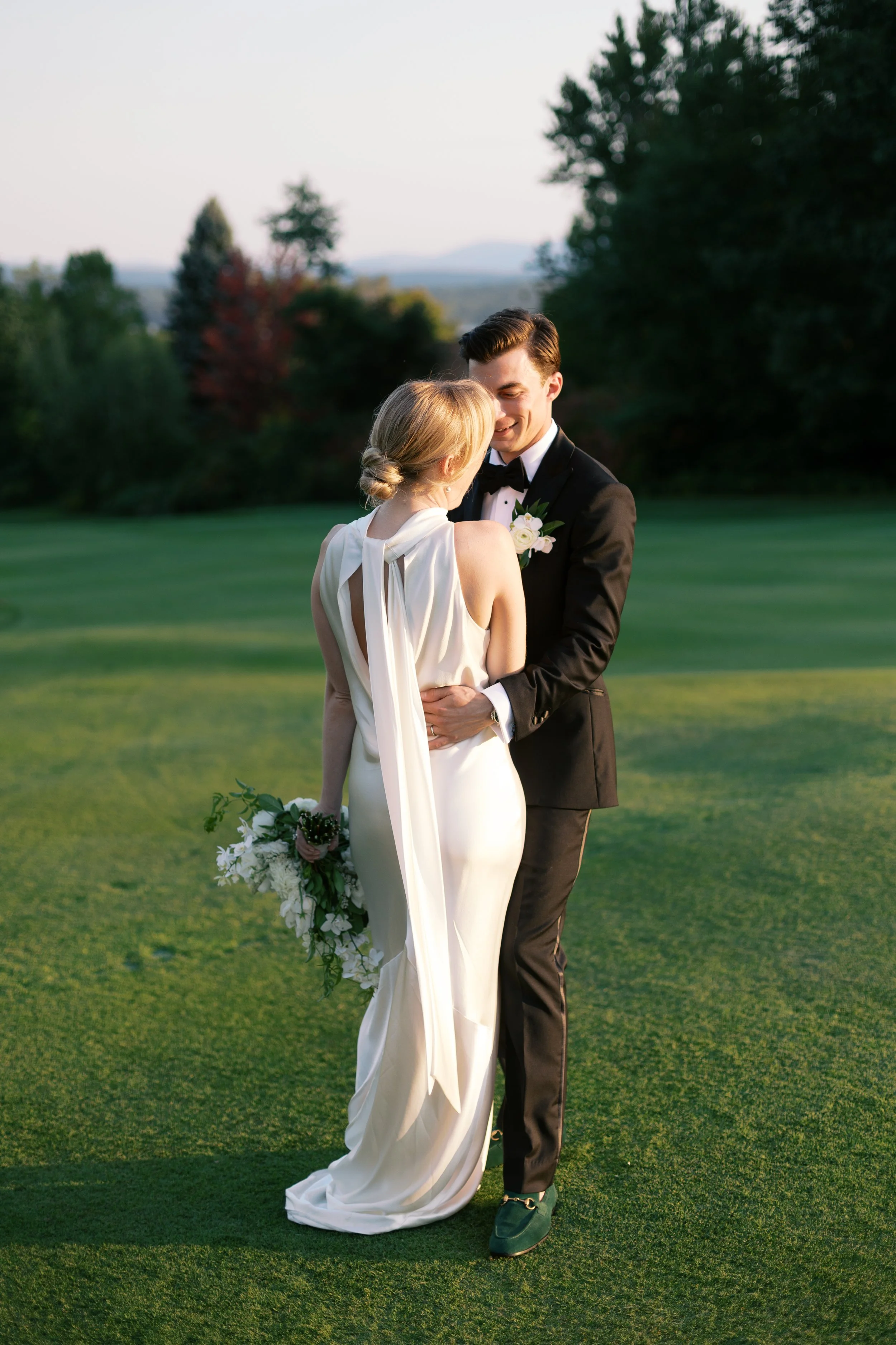 A bride and groom standing closely on a golf course during sunset, embracing each other with the bride holding a bouquet of flowers, both smiling.