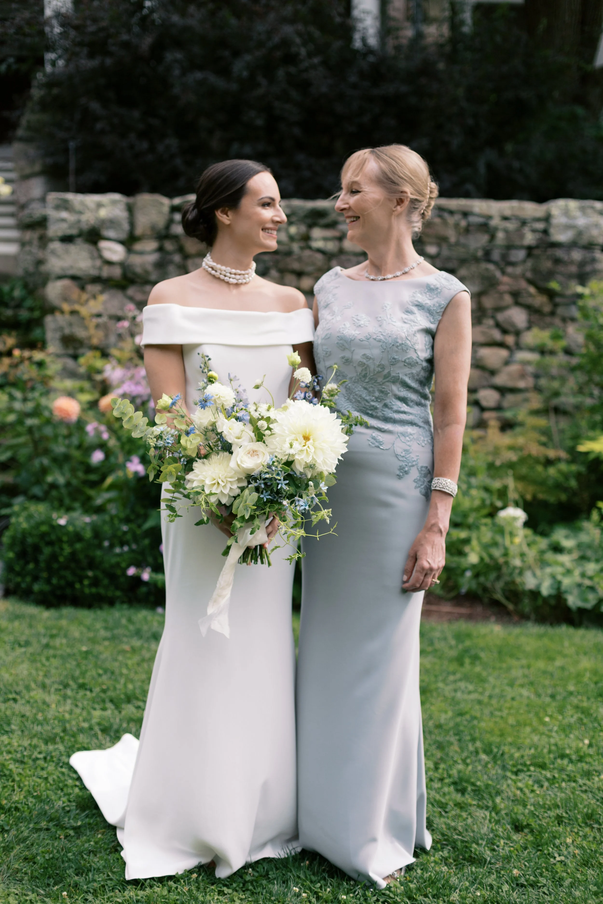 A bride and an older woman, possibly her mother, smiling at each other outdoors. The bride is holding a bouquet of white and green flowers, and they are standing on a grassy area with plants and a stone wall in the background.