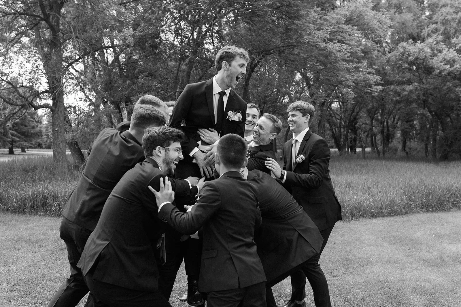 A group of men in suits lifting a man in a tuxedo in the air during an outdoor celebration, smiling and laughing.