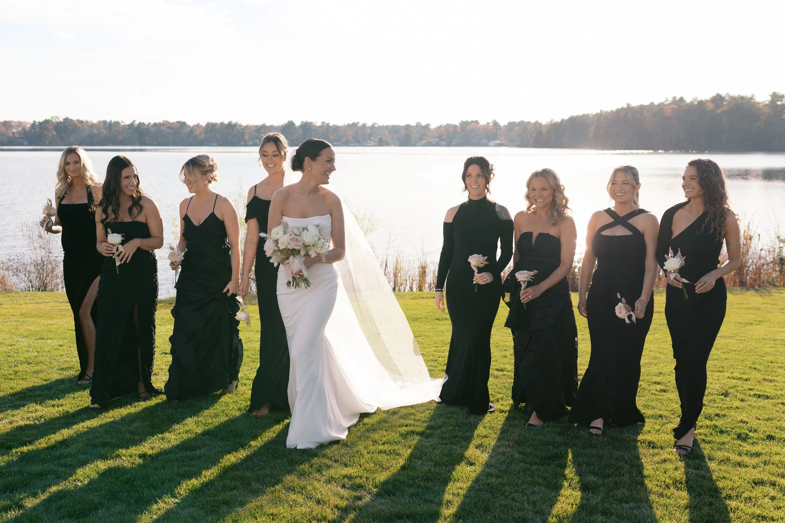 Bride in a white wedding dress holding a bouquet walking with nine bridesmaids in black dresses by a lakeside.