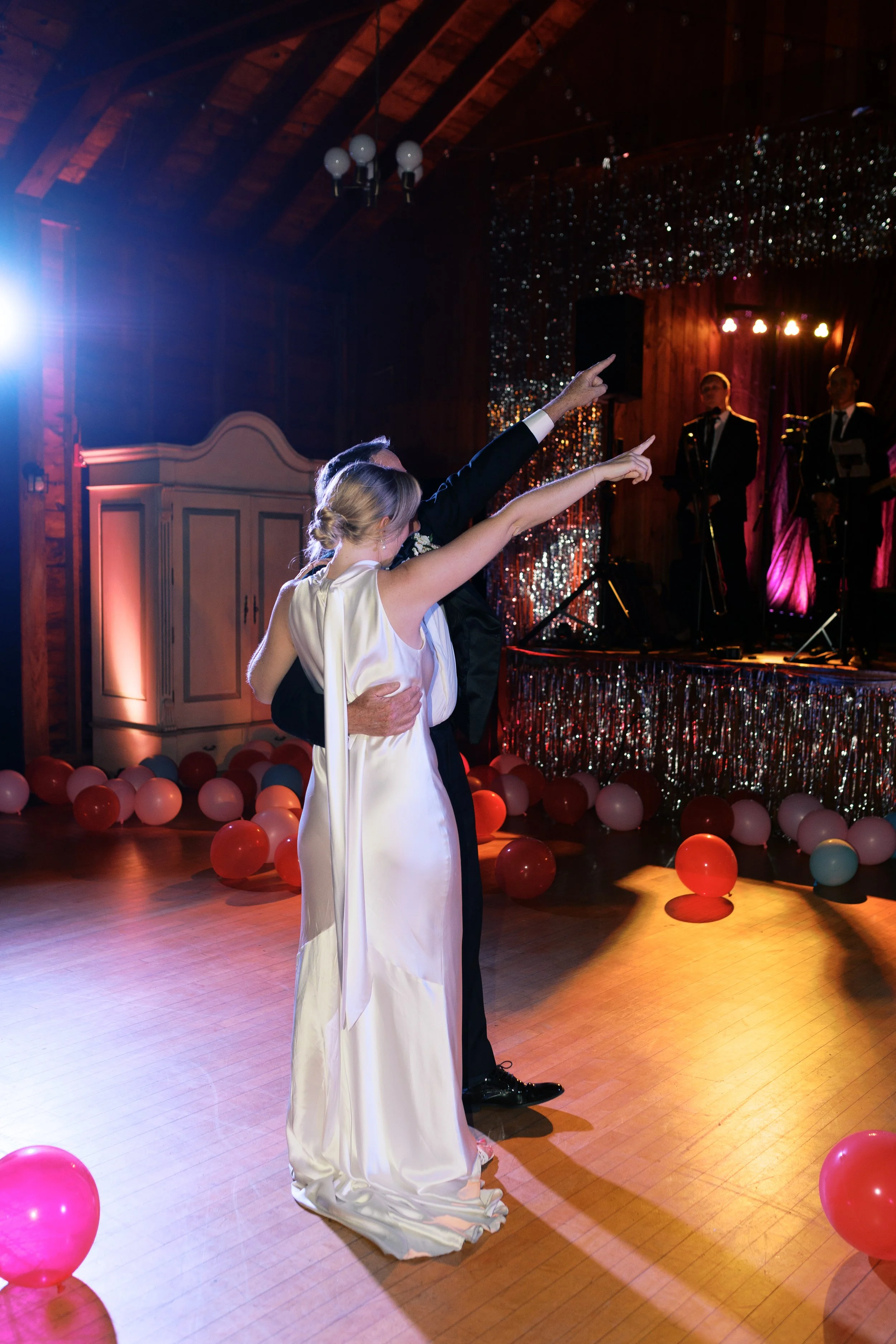 A bride and groom dancing at their wedding reception, surrounded by balloons, with band members performing on stage in the background.