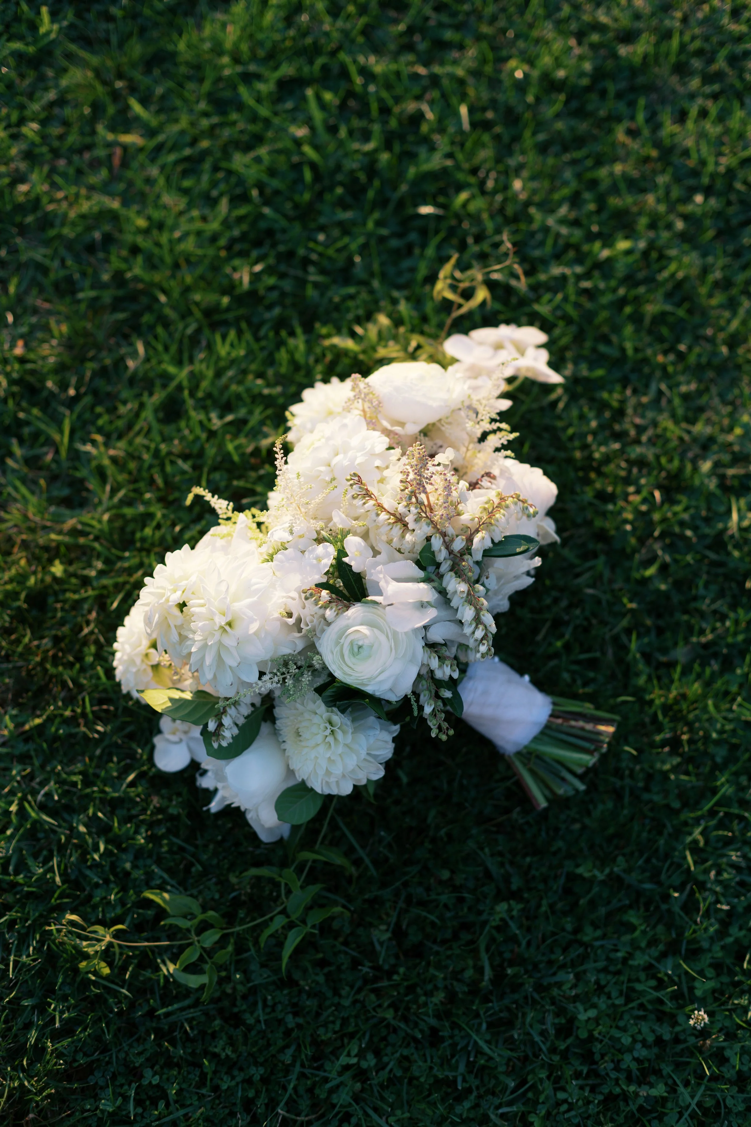 White wedding bouquet with roses, dahlias, and greenery lying on green grass