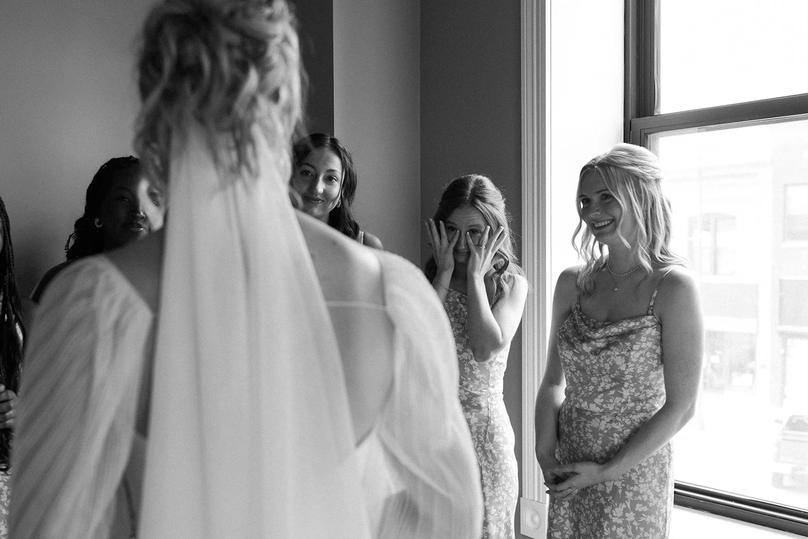 A bride in a wedding dress is surrounded by her bridesmaids, who are reacting emotionally to her, with one covering her face and others smiling, near a large window in a bright room.