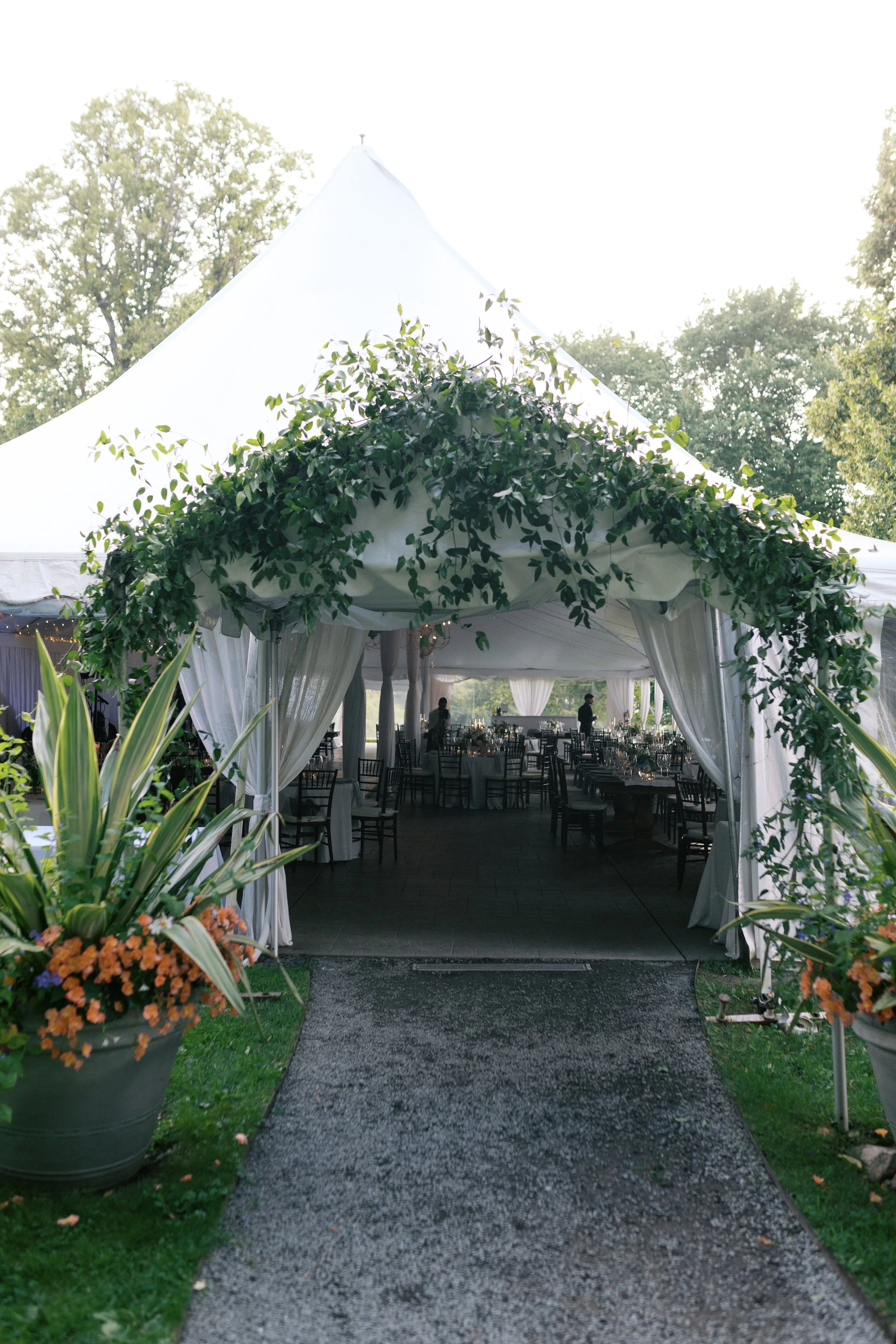 Wedding reception tent with decorated entrance, surrounded by plants and flowers, set up outdoors with tables and chairs inside.
