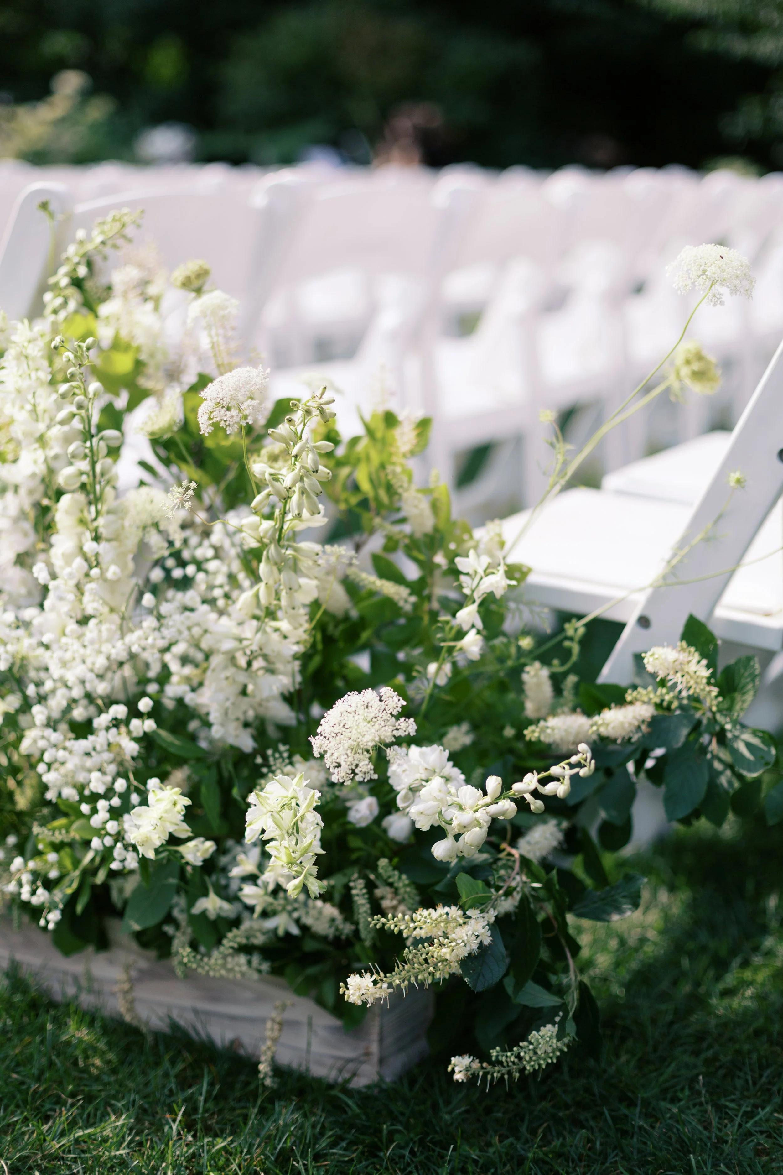 White floral arrangement with various flowers and greenery, placed outdoors on the grass near white chairs for a wedding or event.