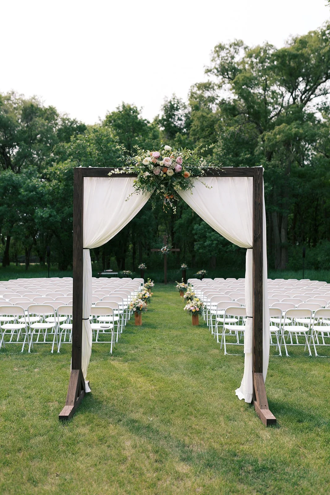 Outdoor wedding setup with white chairs arranged in two rows on either side of a grassy aisle, decorated with flower arrangements, and an arch draped with white curtains and a floral arrangement at the top, set against a backdrop of green trees.