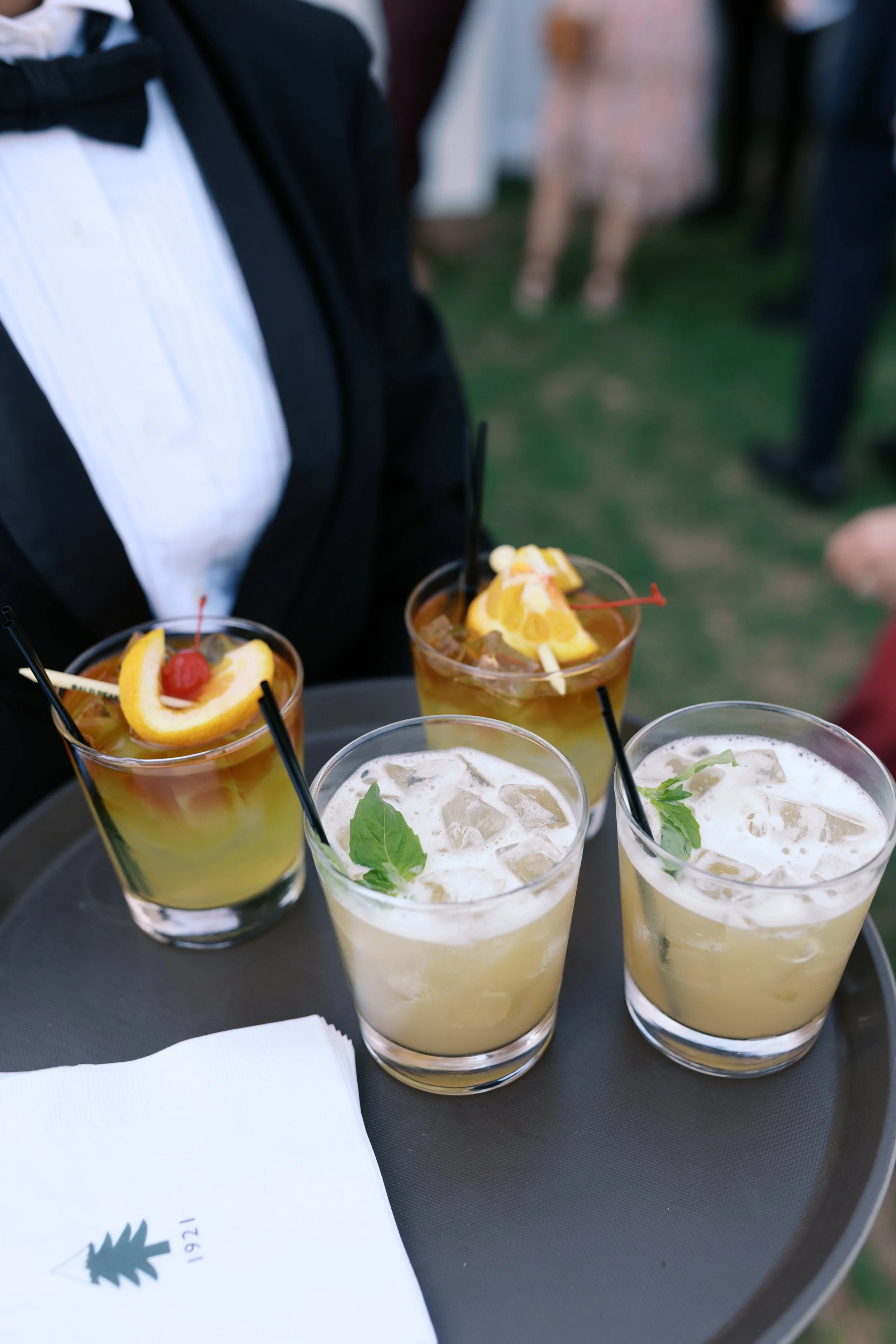 Four cocktails with ice and garnishes on a black tray, being held by a person in formal attire at an outdoor event.
