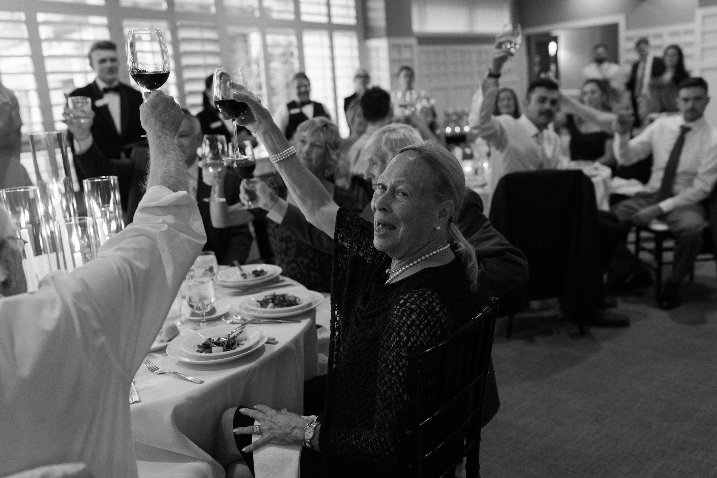 Black and white photo of a woman raising a glass of wine at a formal dinner, surrounded by other guests toasting, in a well-lit restaurant or banquet hall.