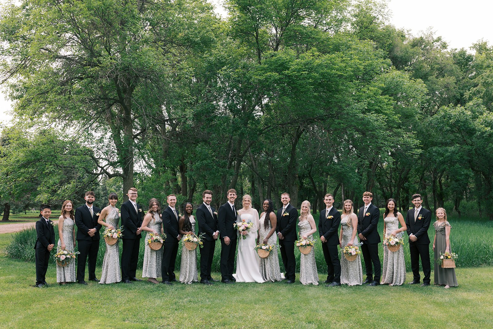 A wedding party of 17 people standing outdoors on a grassy area with trees in the background. The bride and groom are in the center, surrounded by bridesmaids and groomsmen. The women are wearing matching long, patterned dresses, and the men are in d