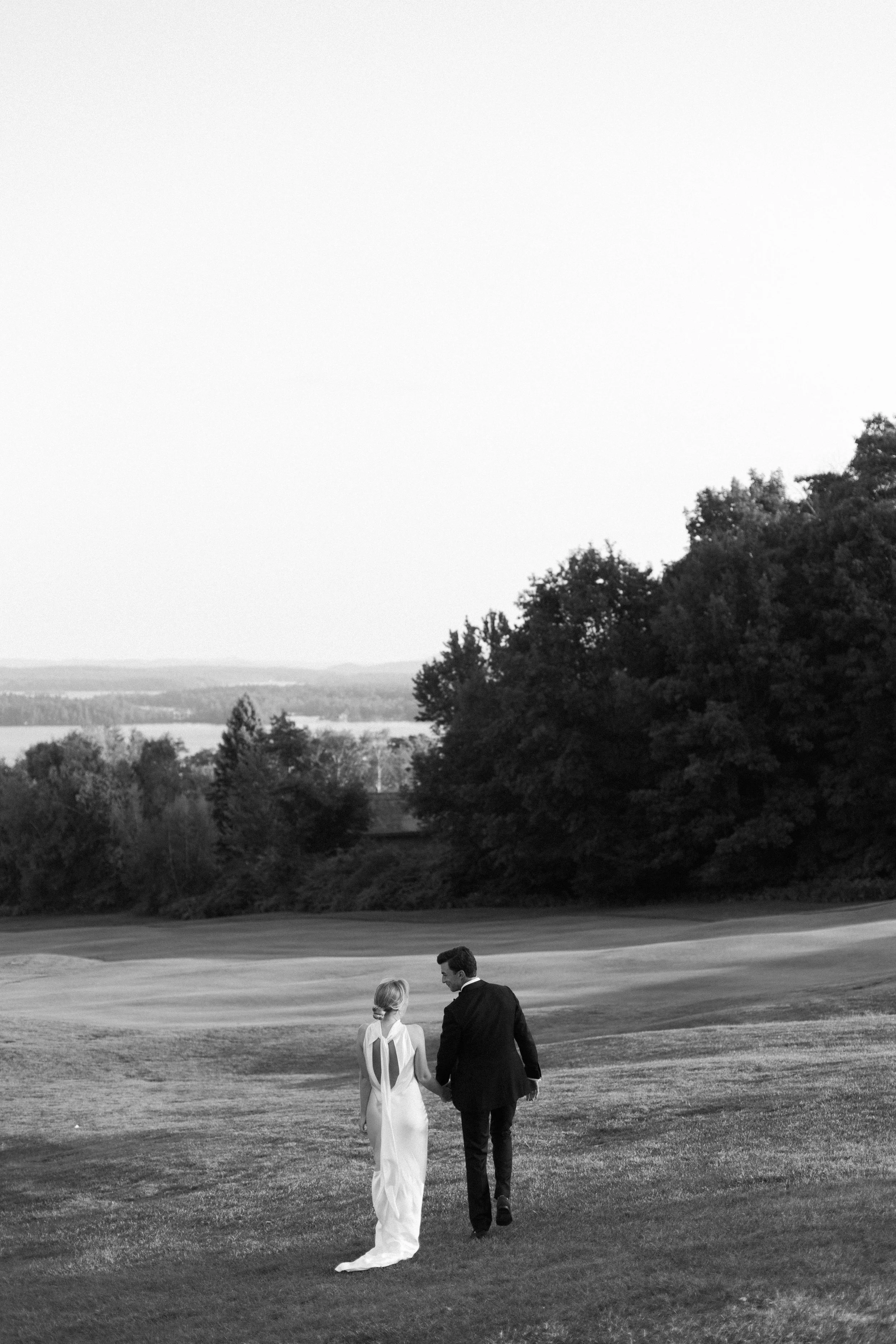 A bride and groom walking hand in hand on a grassy field, with trees and hills in the distance, in black and white.