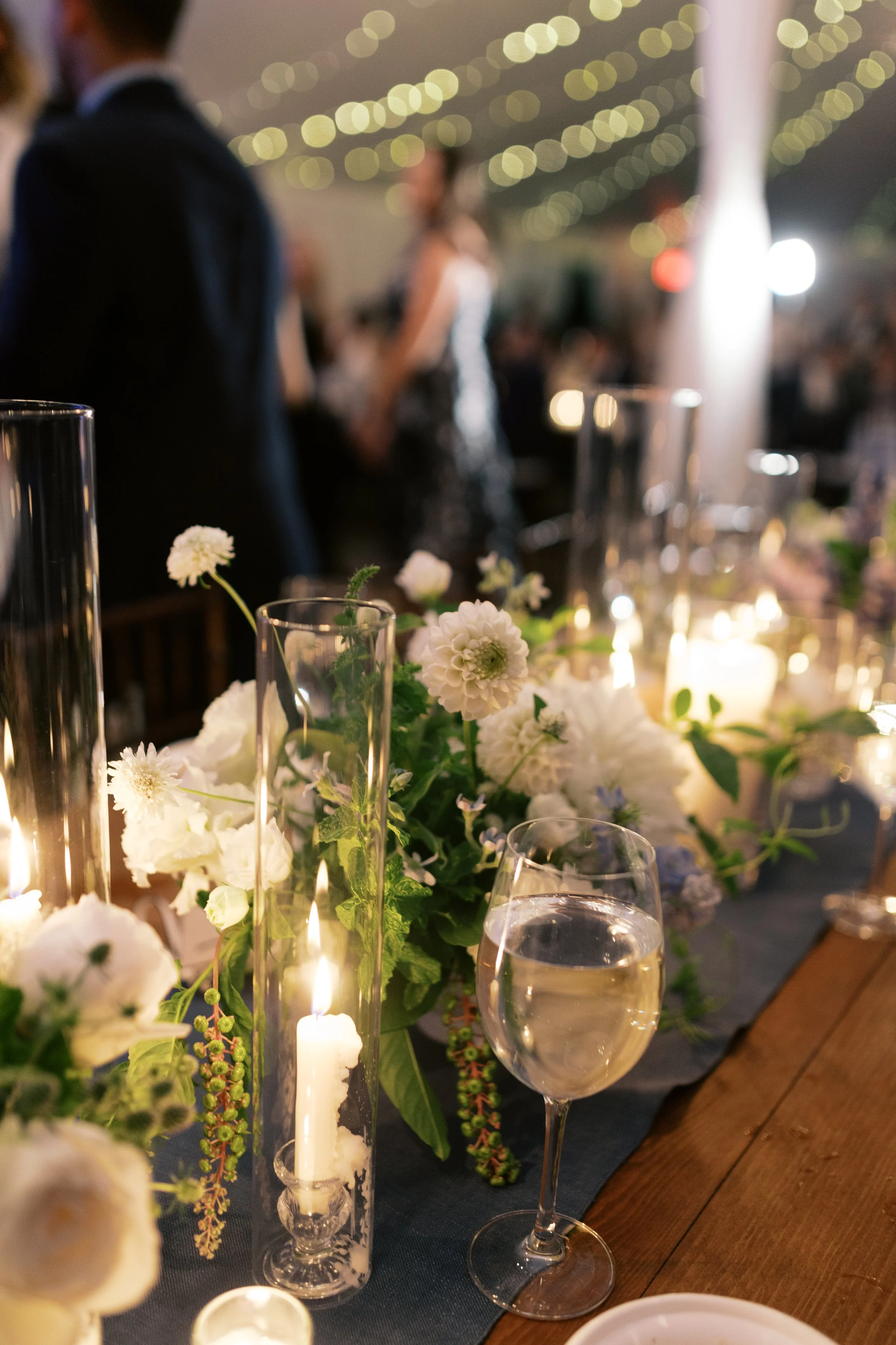 Elegant dinner table with white floral centerpiece, lit candles in glass holders, and a glass of white wine, with blurred guests and string lights in the background.