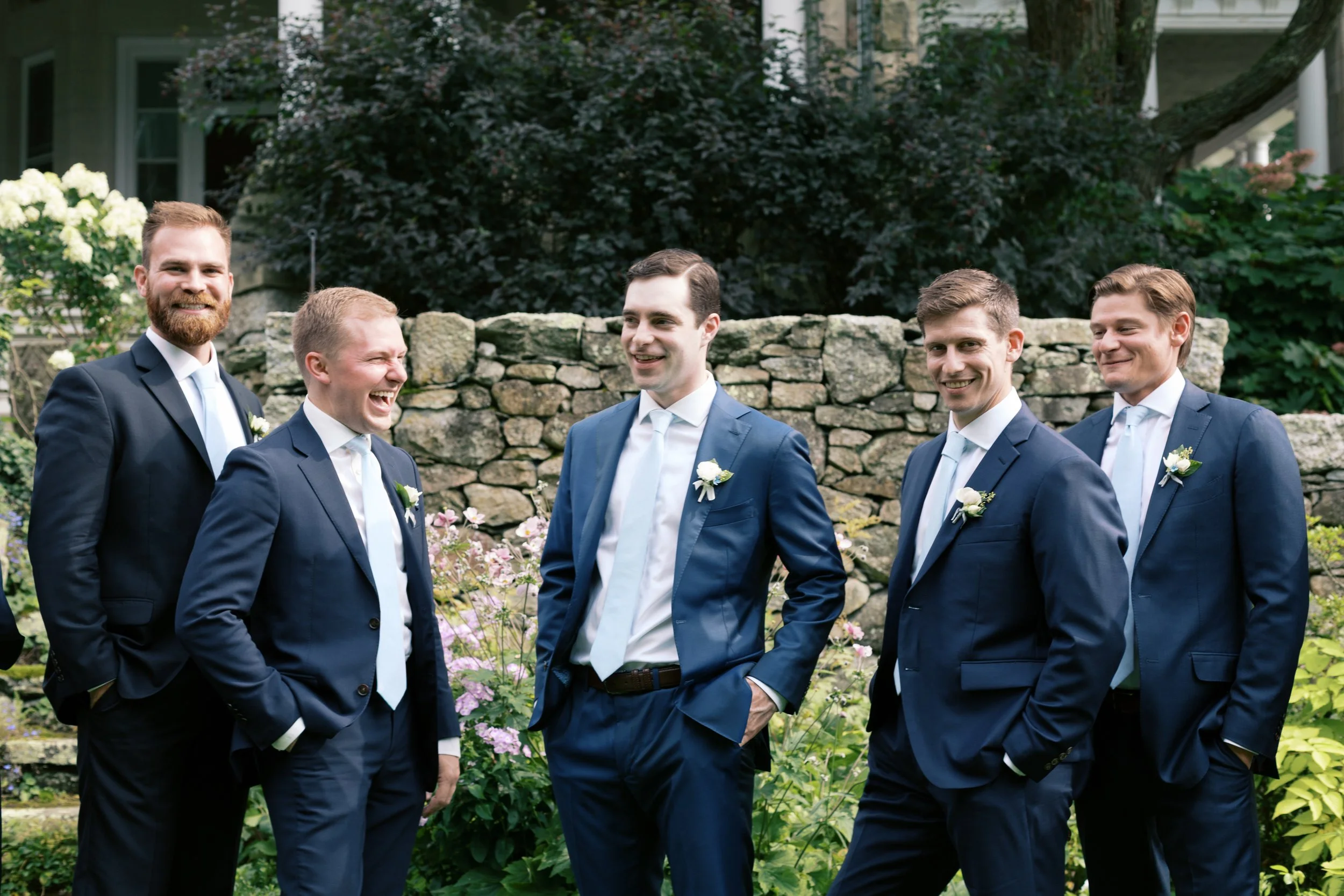 Six men in blue suits and white ties standing outdoors in a garden, smiling, with a stone wall and greenery in the background.