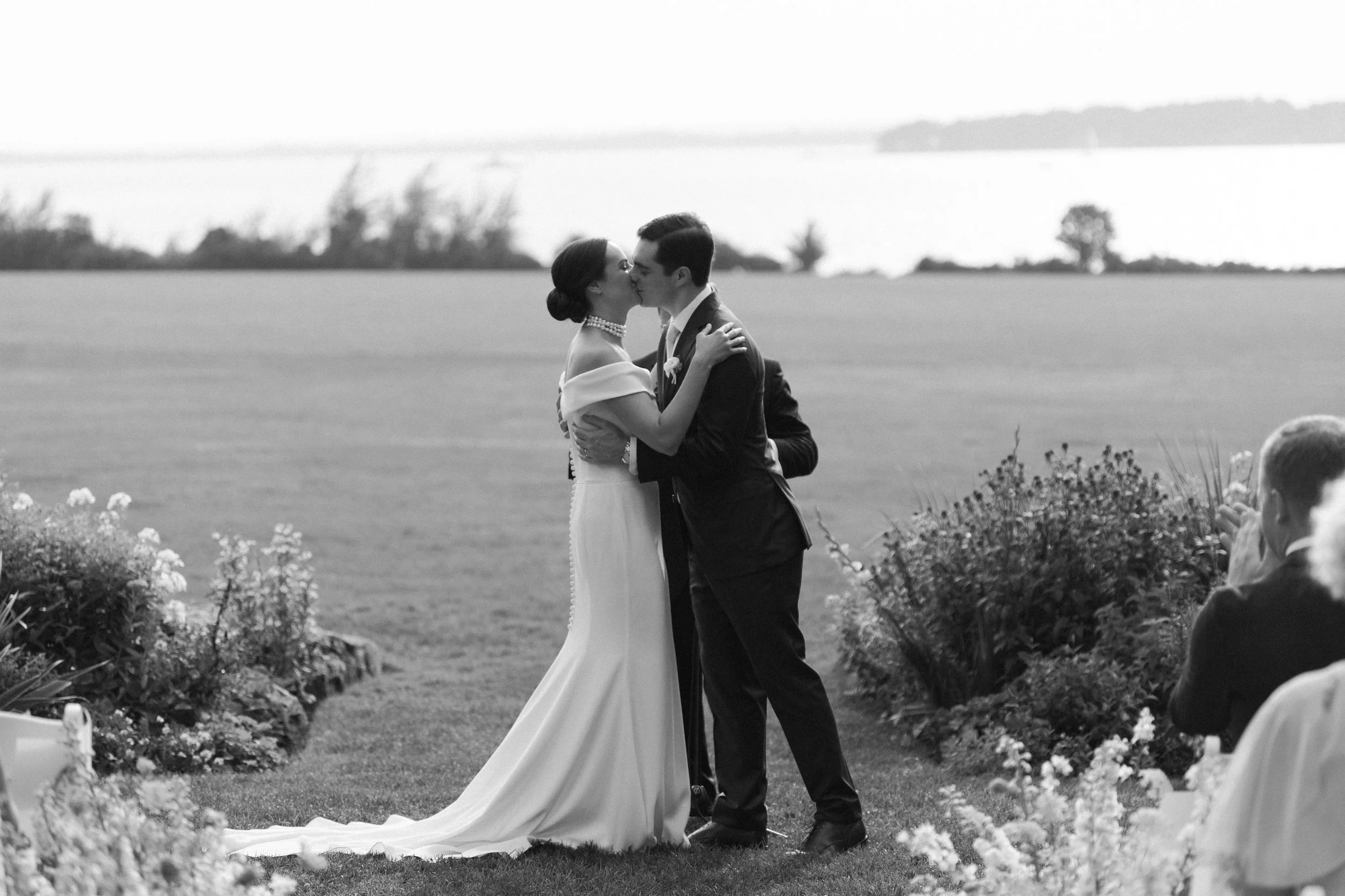 A black and white photo of a couple kissing during their wedding ceremony outdoors, with wedding guests visible in the foreground and a scenic water body in the background.