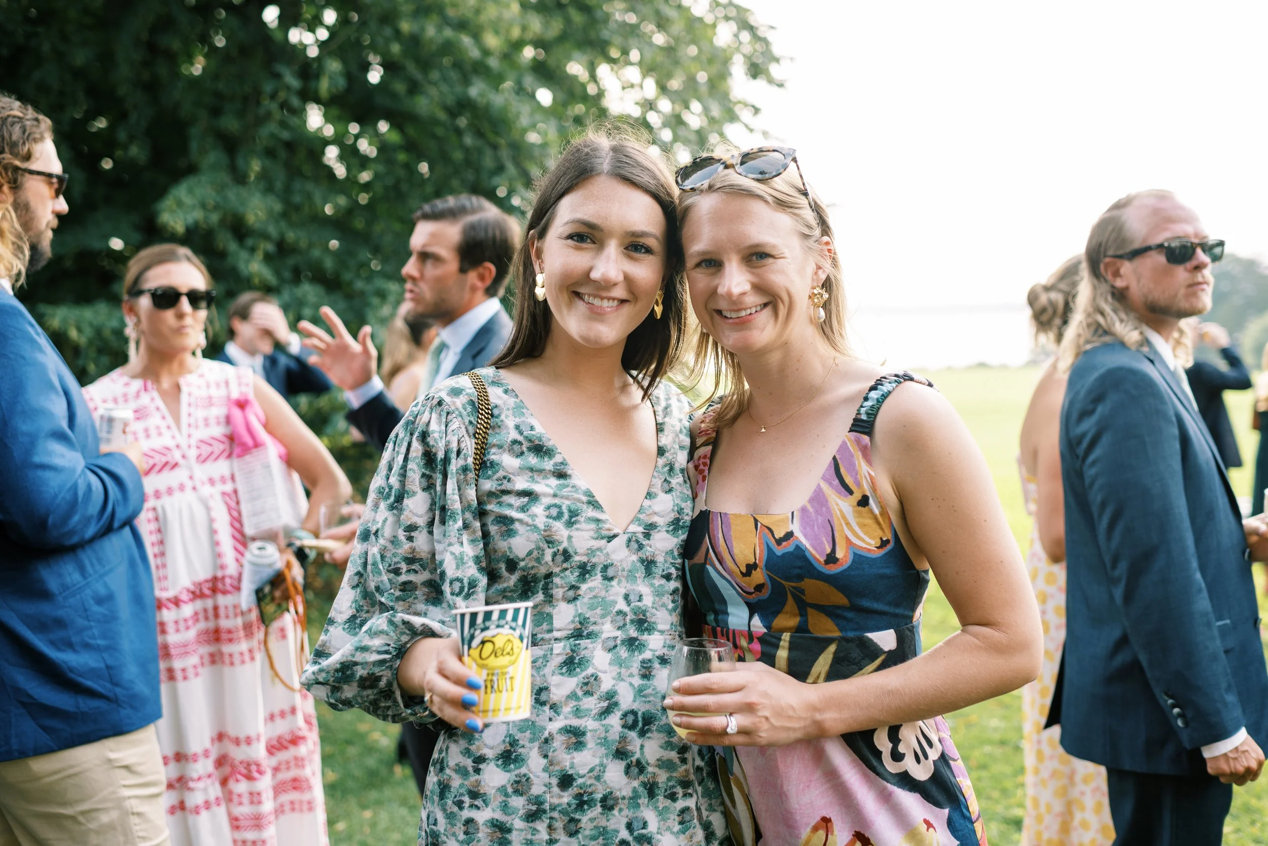 Two women smiling and posing for a photo at an outdoor social gathering, with other guests in the background.