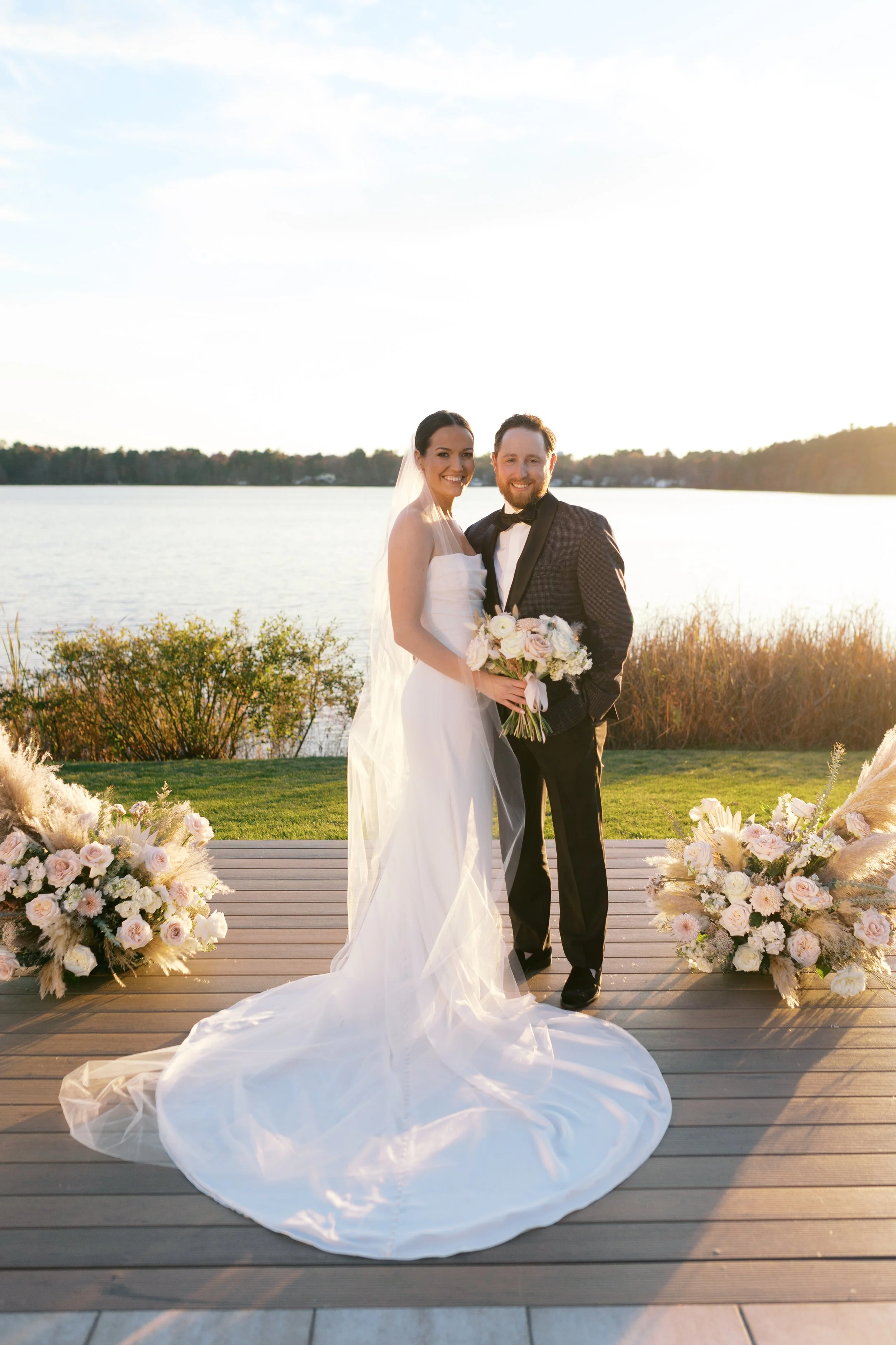 A bride and groom standing on a wooden platform outdoors near a body of water, holding a bouquet of flowers, with floral arrangements on each side, during sunset.