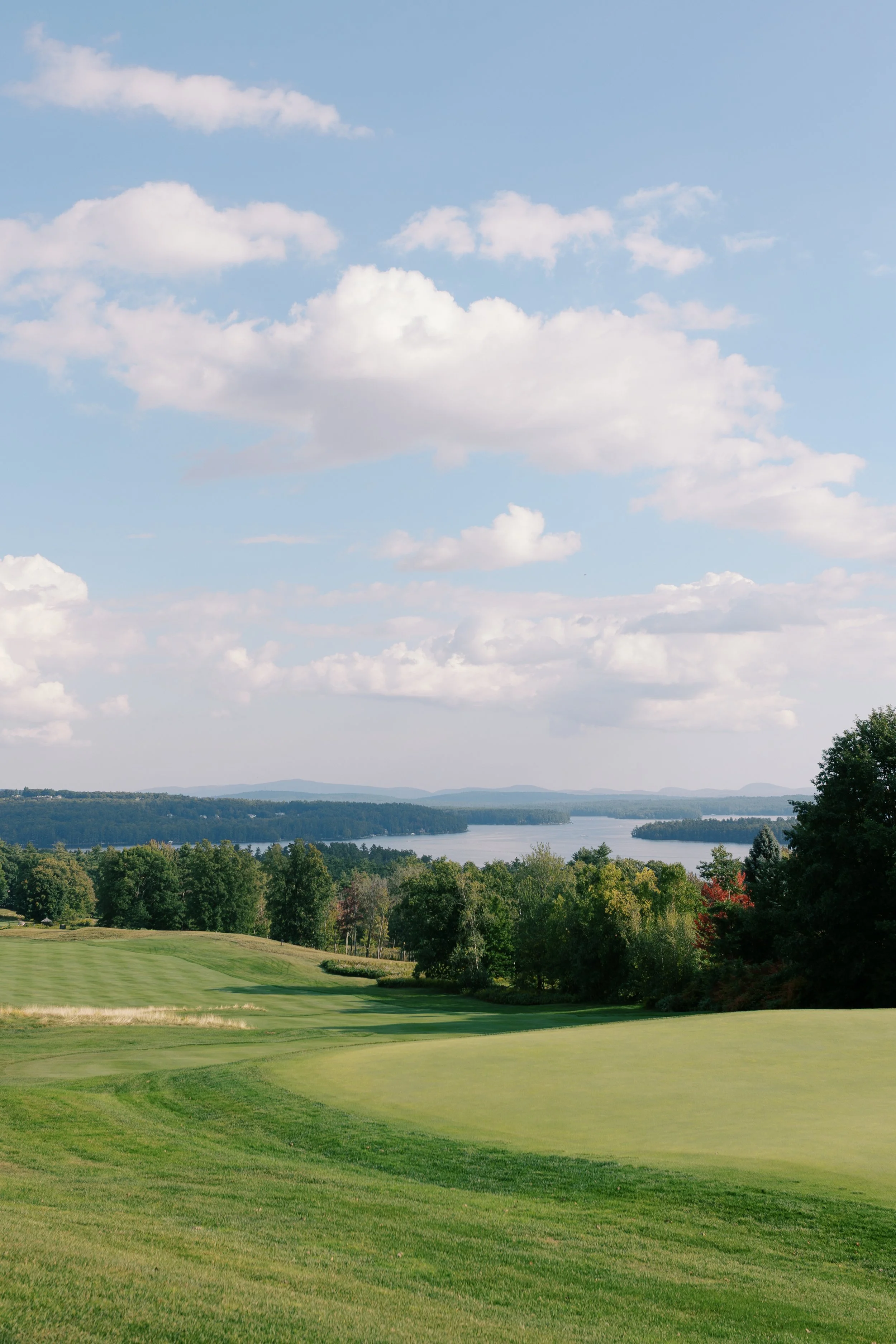 A scenic view of a golf course with well-manicured greens, surrounded by trees, with a lake and distant hills under a partly cloudy sky.