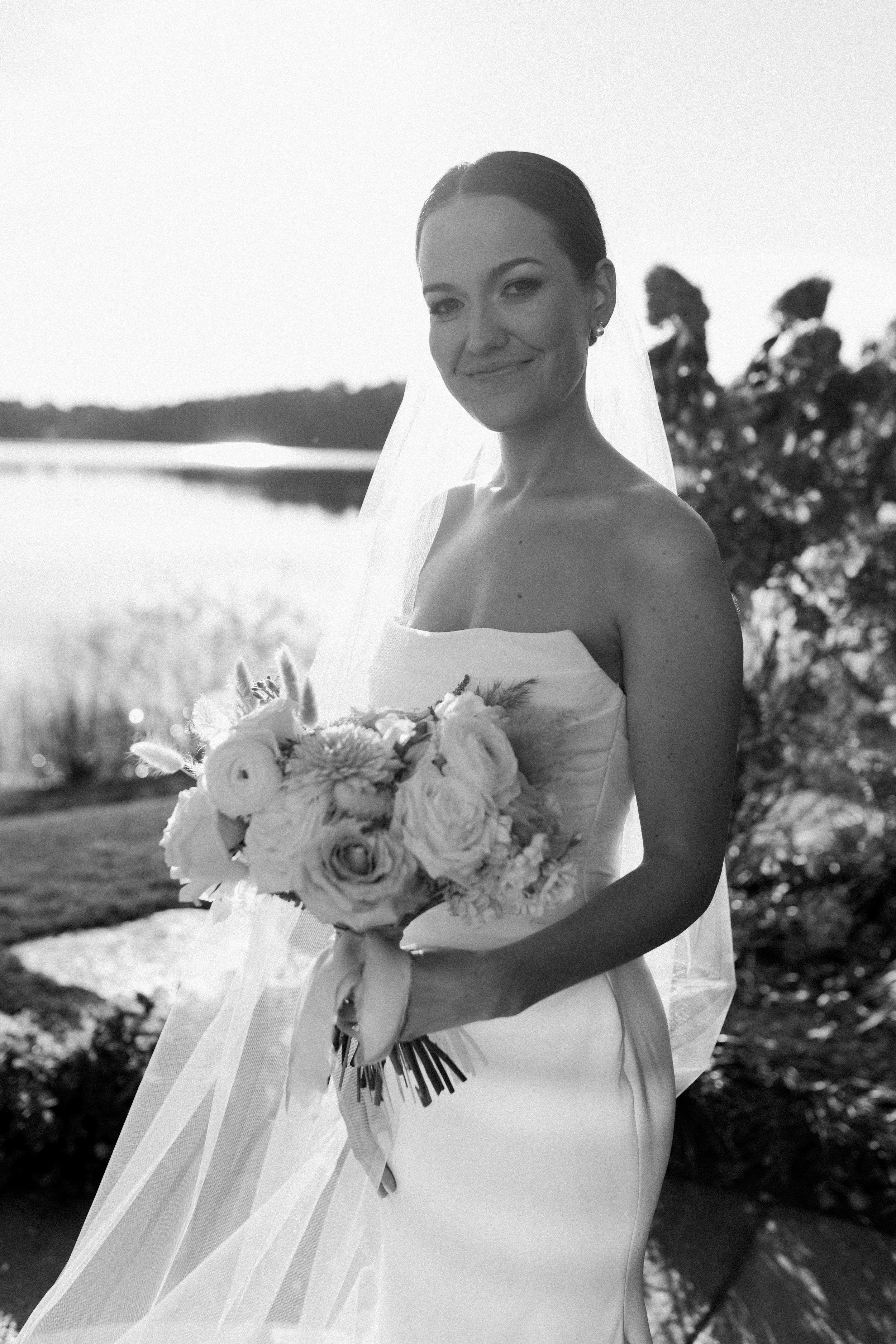 A smiling bride holding a bouquet of flowers outdoors near a body of water, with trees in the background.