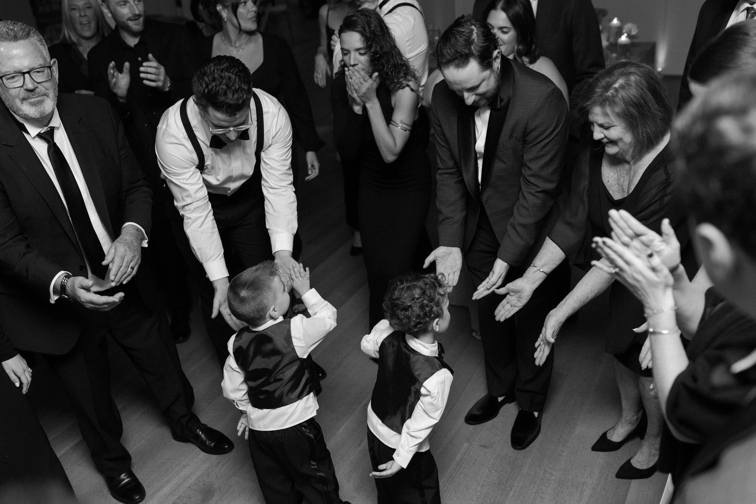 People dancing and celebrating at a party, including children and adults, with two boys and adults. The children are giving high-fives to the adults, who are clapping and smiling.