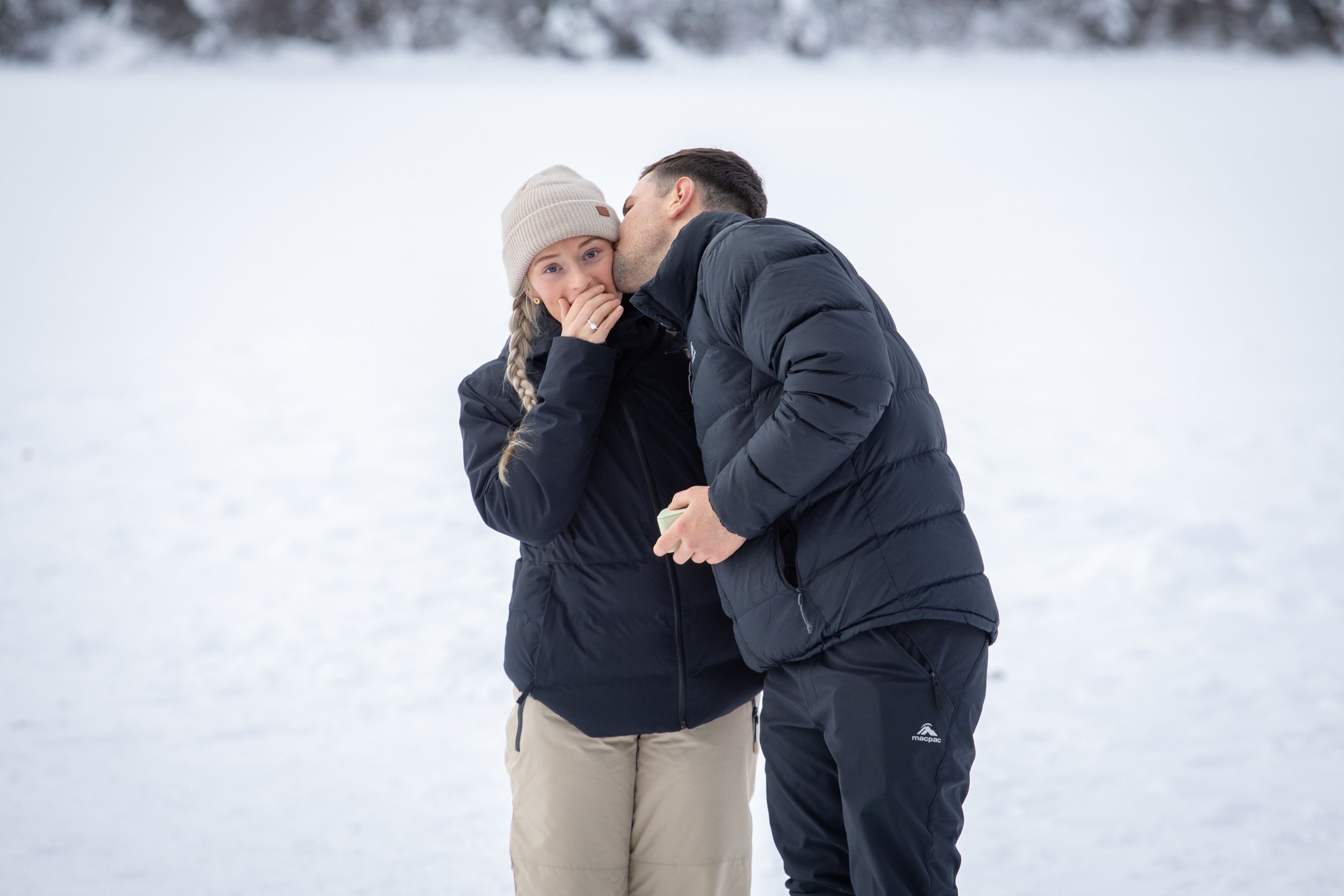 A man is kissing a woman on the cheek while she looks surprised and covers her mouth with her hand in a snowy outdoor setting. Photo by Megan Posein Photography