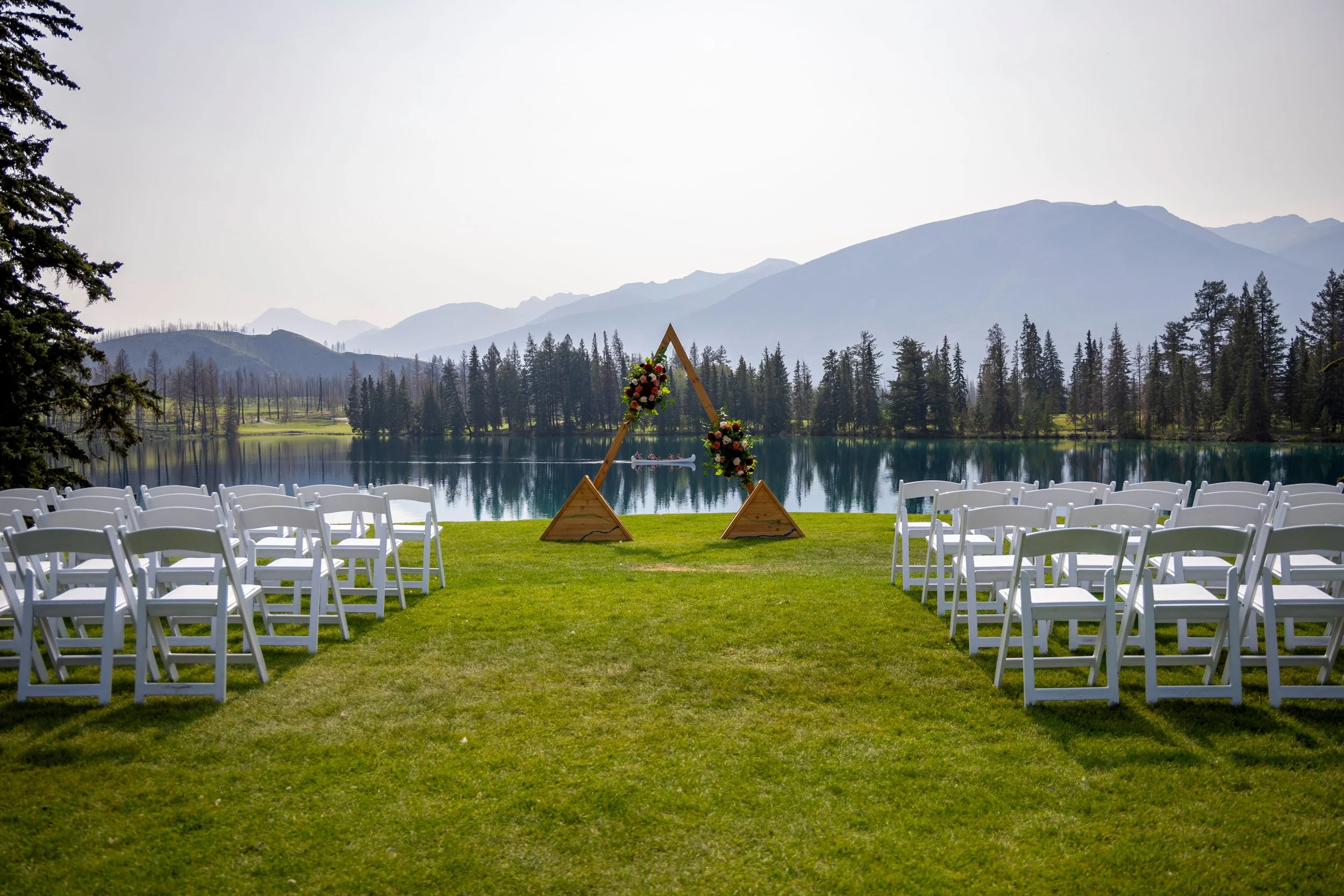 Outdoor wedding ceremony at the Jasper Park Lodge setup by a lake with mountains in the background, featuring a wooden triangular arch decorated with flowers, white chairs arranged on green grass. Photo by Megan Posein Photography