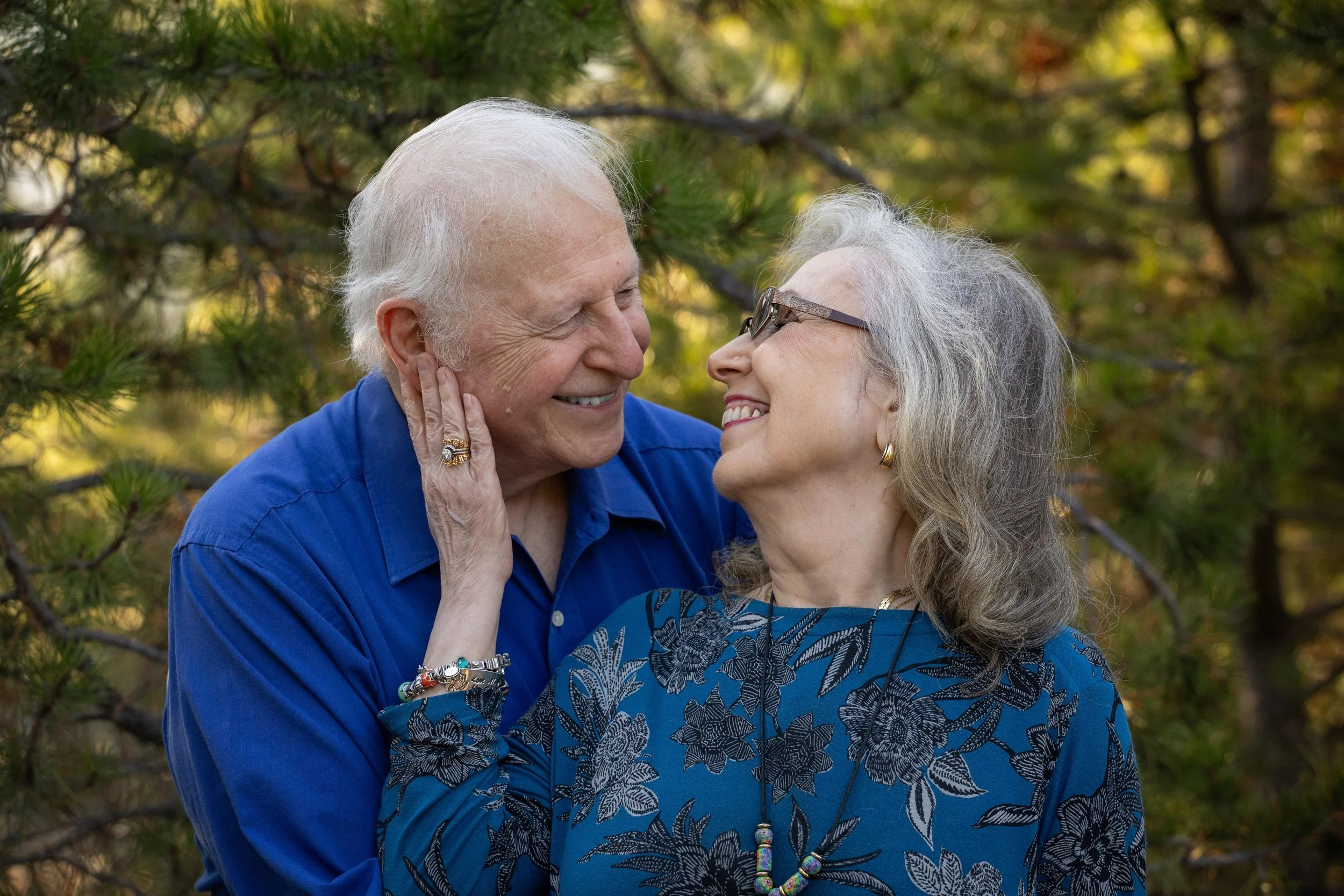 An elderly man and woman smiling and gazing at each other outdoors, surrounded by autumn foliage. Photo by Megan Posein Photography