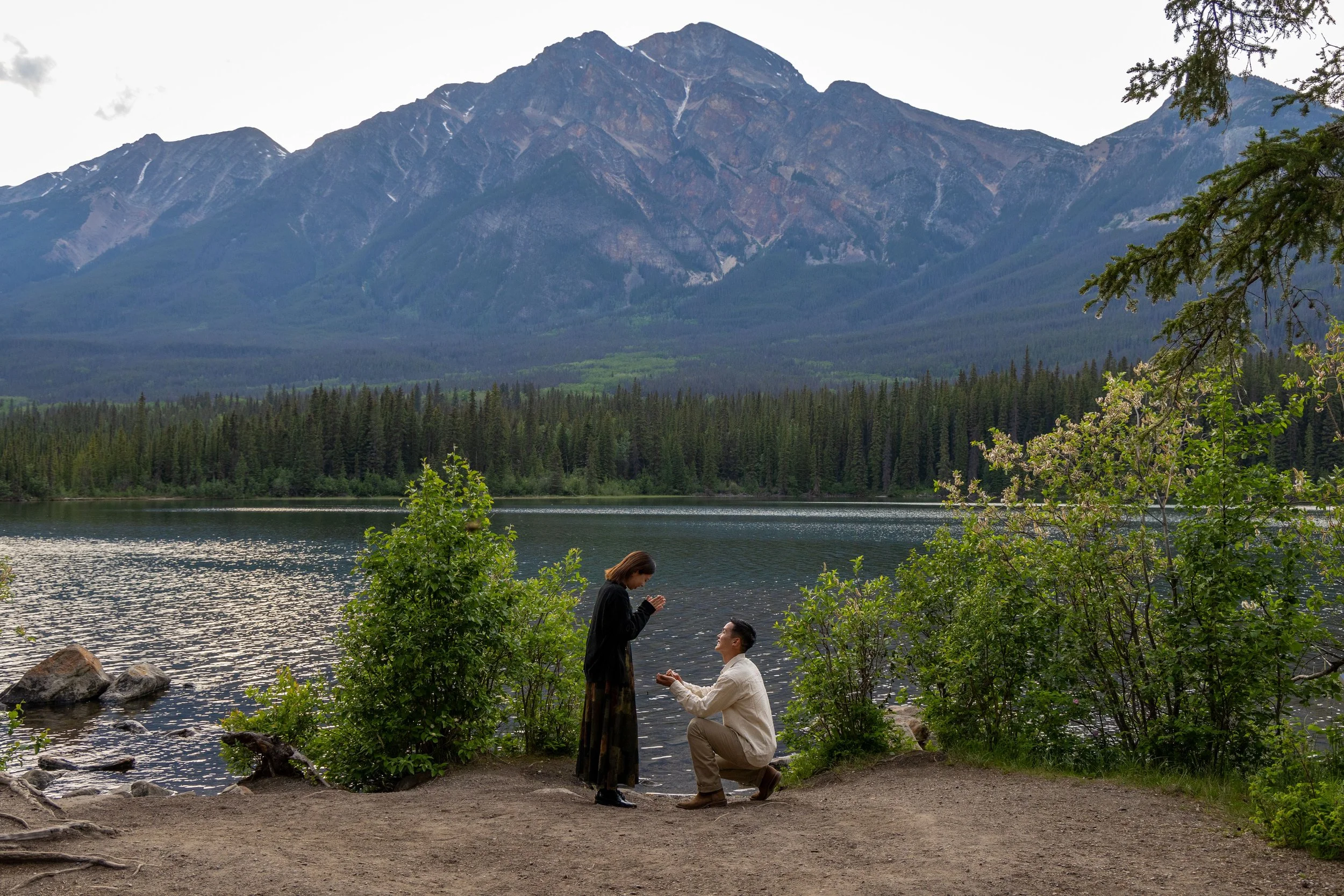 A man proposes marriage to a woman by a lakeside with mountains and pine trees in the background. Photo by Megan Posein Photography