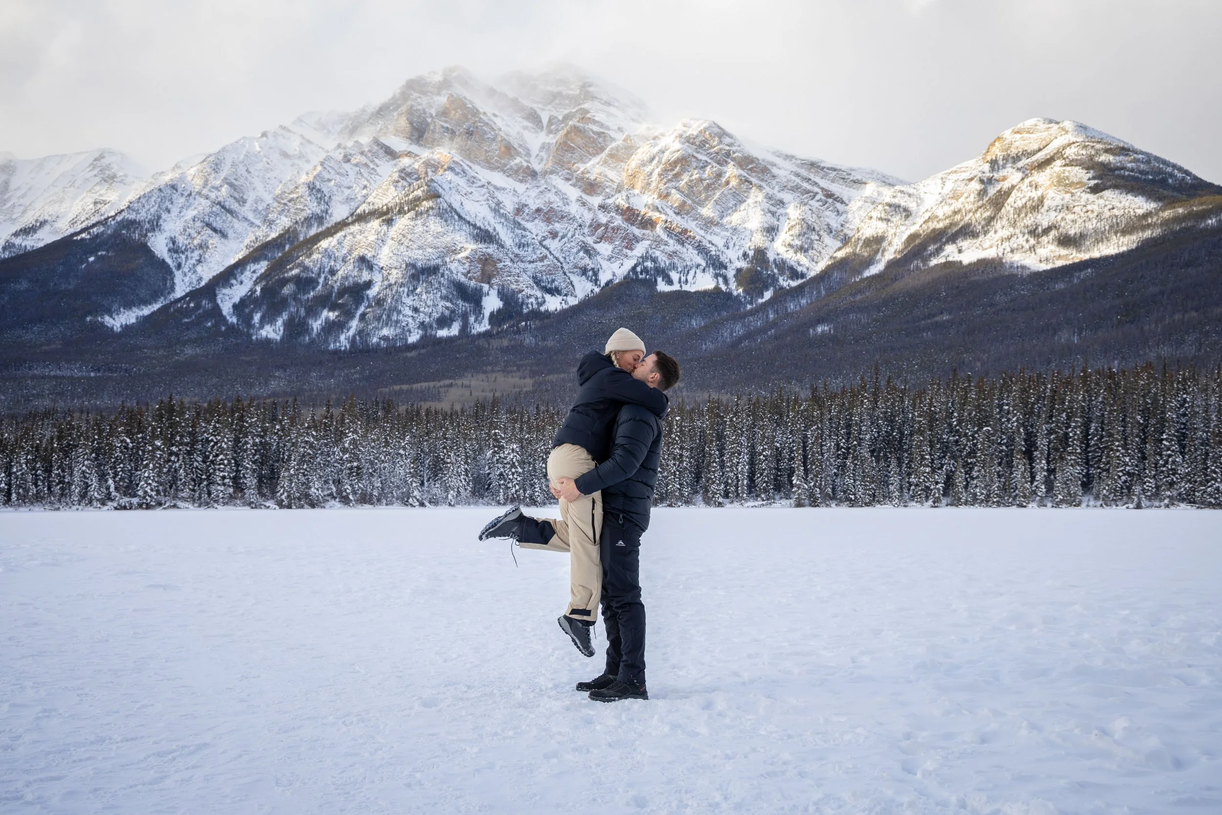 A couple in winter clothing embracing in a snowy landscape with snow-covered mountains and a forest of pine trees in the background. Photo by Megan Posein Photography