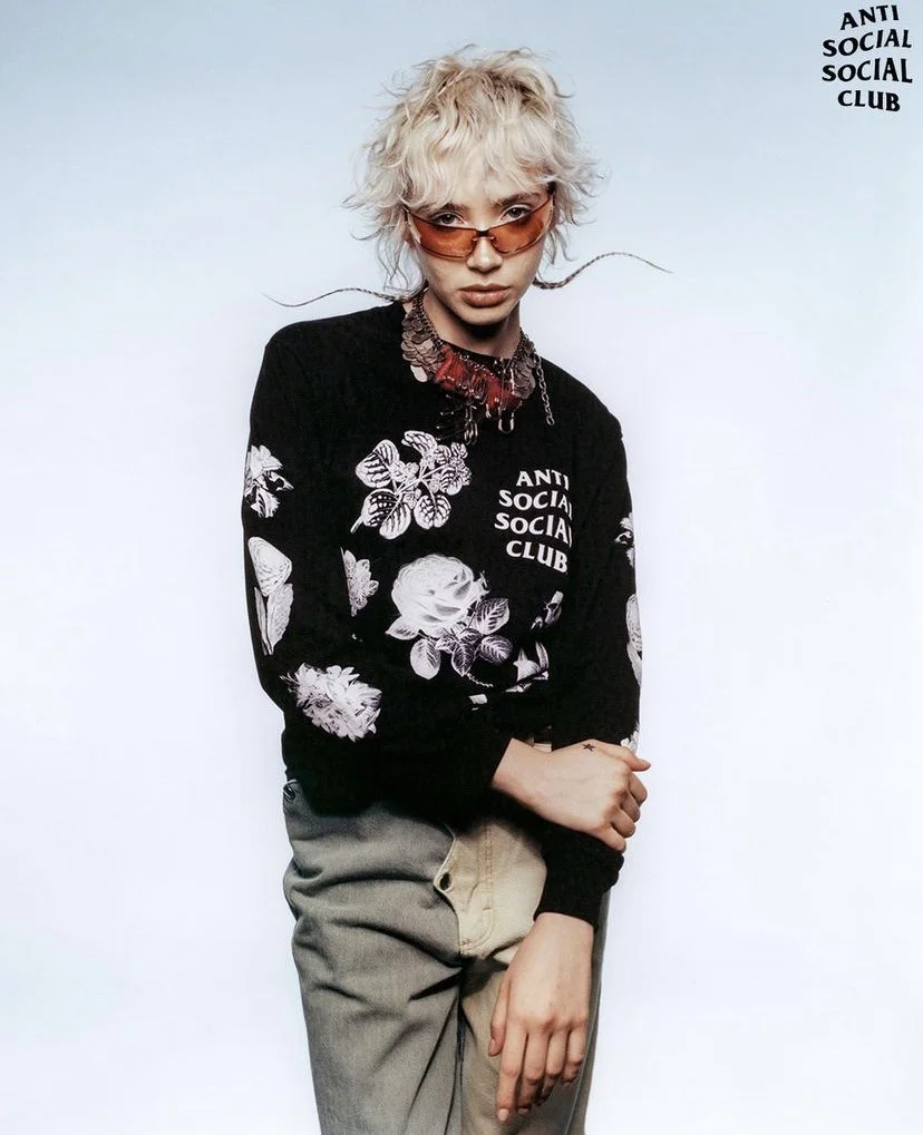 Young person with blonde, curly hair and orange-tinted glasses wearing a black sweatshirt with white floral and butterfly designs and the words 'Anti Social Social Club' against a light background.