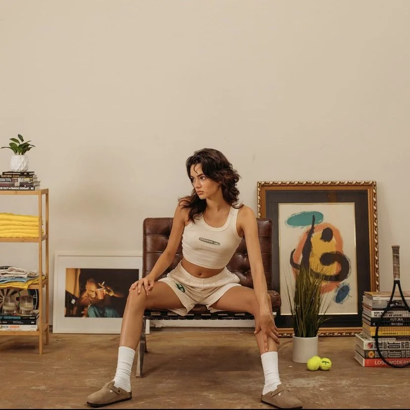Young woman sitting on a vintage chair in a room with artworks and plants, wearing casual athletic clothes.