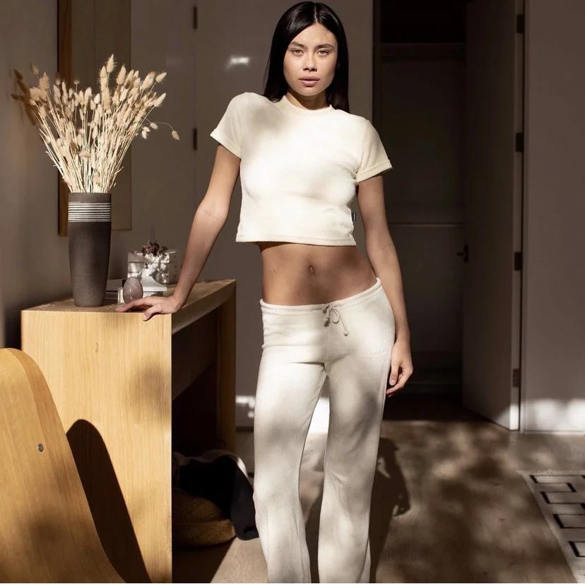 Young woman with long dark hair wearing a light-colored t-shirt and matching sweatpants, standing indoors near a wooden table with a tall vase of dried flowers.