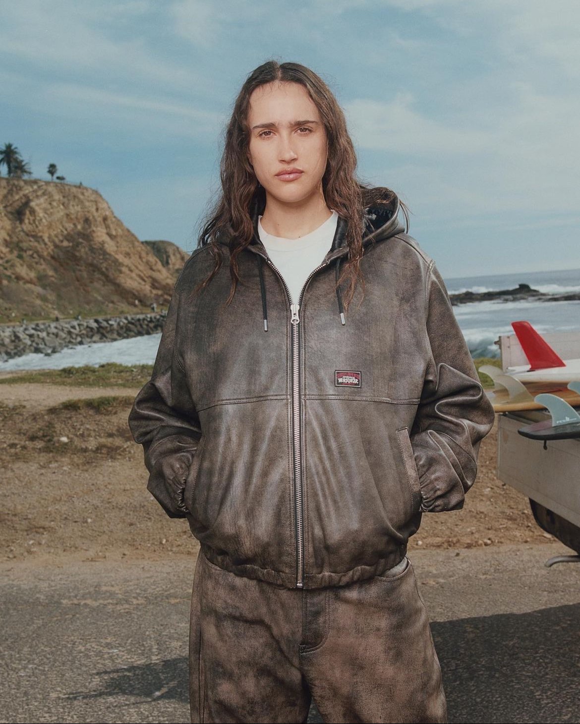 A young woman with long brown wavy hair, standing outdoors on a beach, wearing a brown leather jacket and matching pants, with hands in pockets, against a background of cliffs, ocean, and parked surfboards.