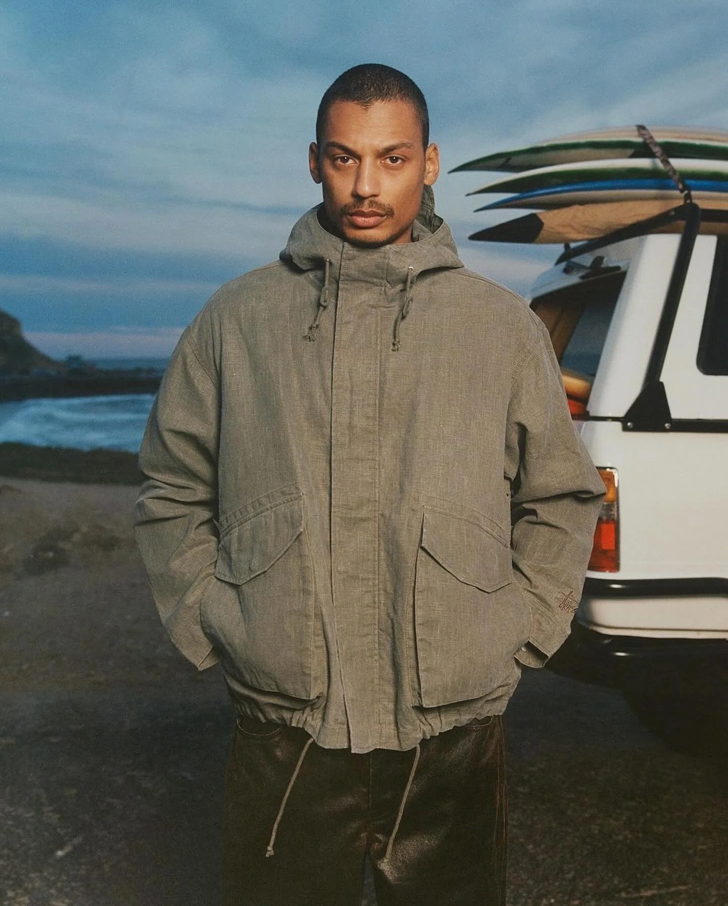 A man standing outdoors in front of a beach and a white van with surfboards on top, wearing a gray jacket with hands in pockets.