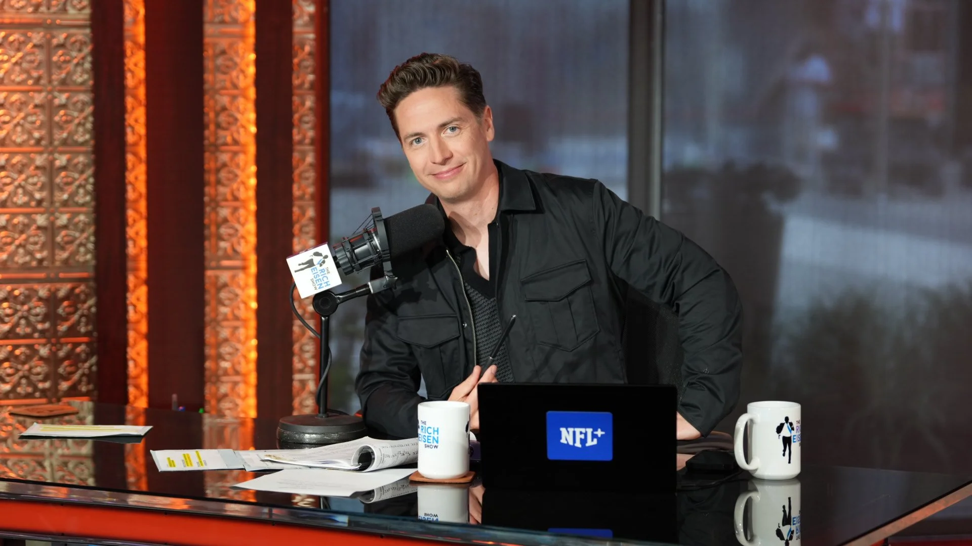 A man with light skin, wearing a black jacket, sitting at a desk with a microphone, a coffee mug, a laptop, and papers. The background includes orange decorative panels and a large window.