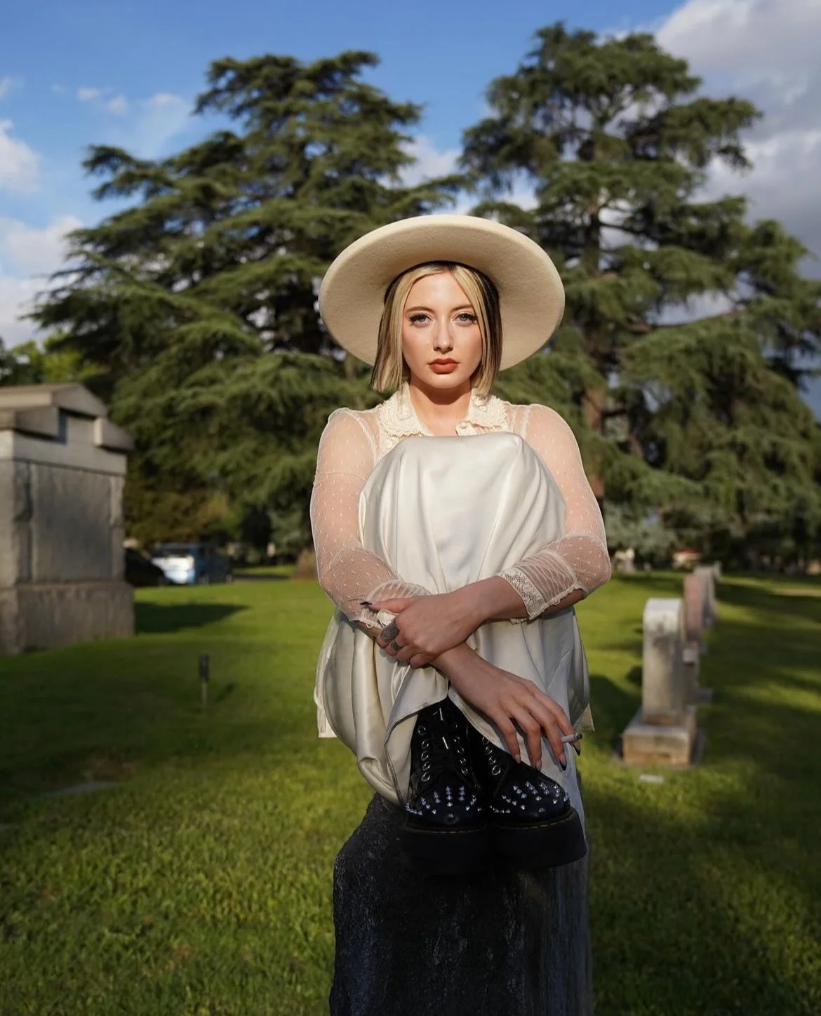 A woman with blonde hair wearing a large hat and a vintage cream-colored dress sitting on a tree stump in a cemetery with granite headstones and lush green grass, trees, and a partly cloudy sky in the background.