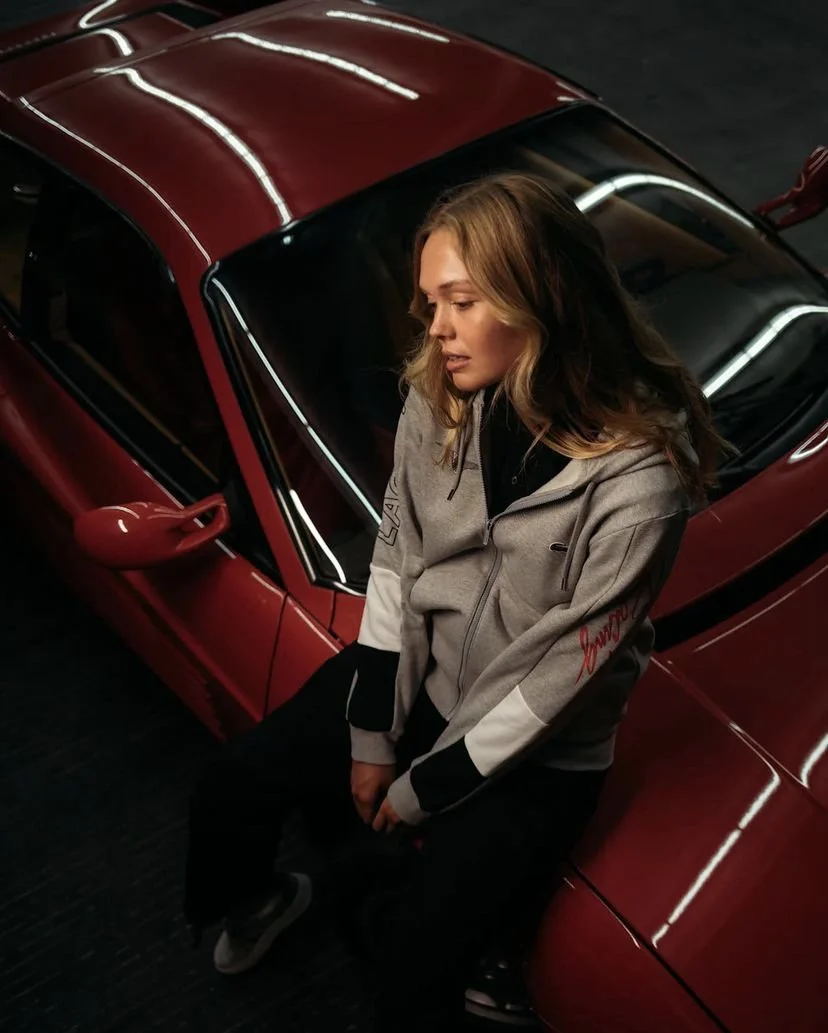 A woman with wavy blonde hair sitting on the floor beside a red sports car, wearing a gray and black jacket.