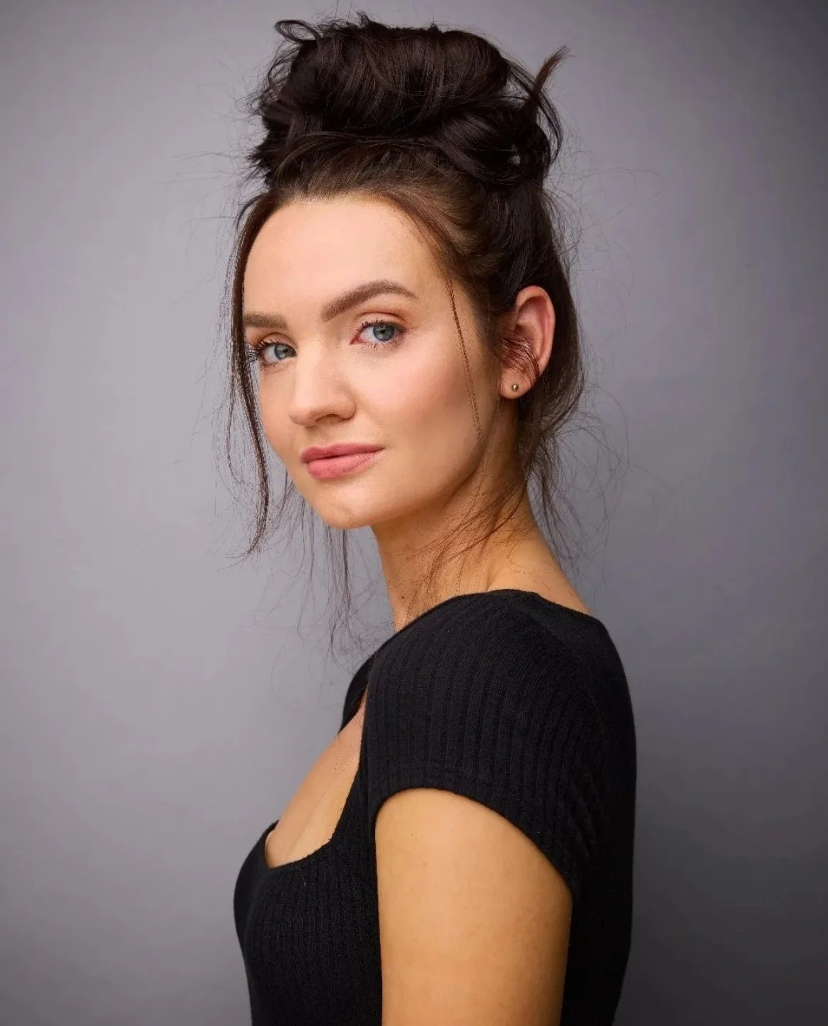 A portrait of a young woman with blue eyes, brown hair styled in an updo, wearing a black top, standing against a gray background.