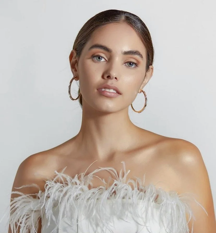 A woman with light skin and brown hair styled in a sleek bun, wearing gold hoop earrings and a white off-shoulder dress with feathered accents.